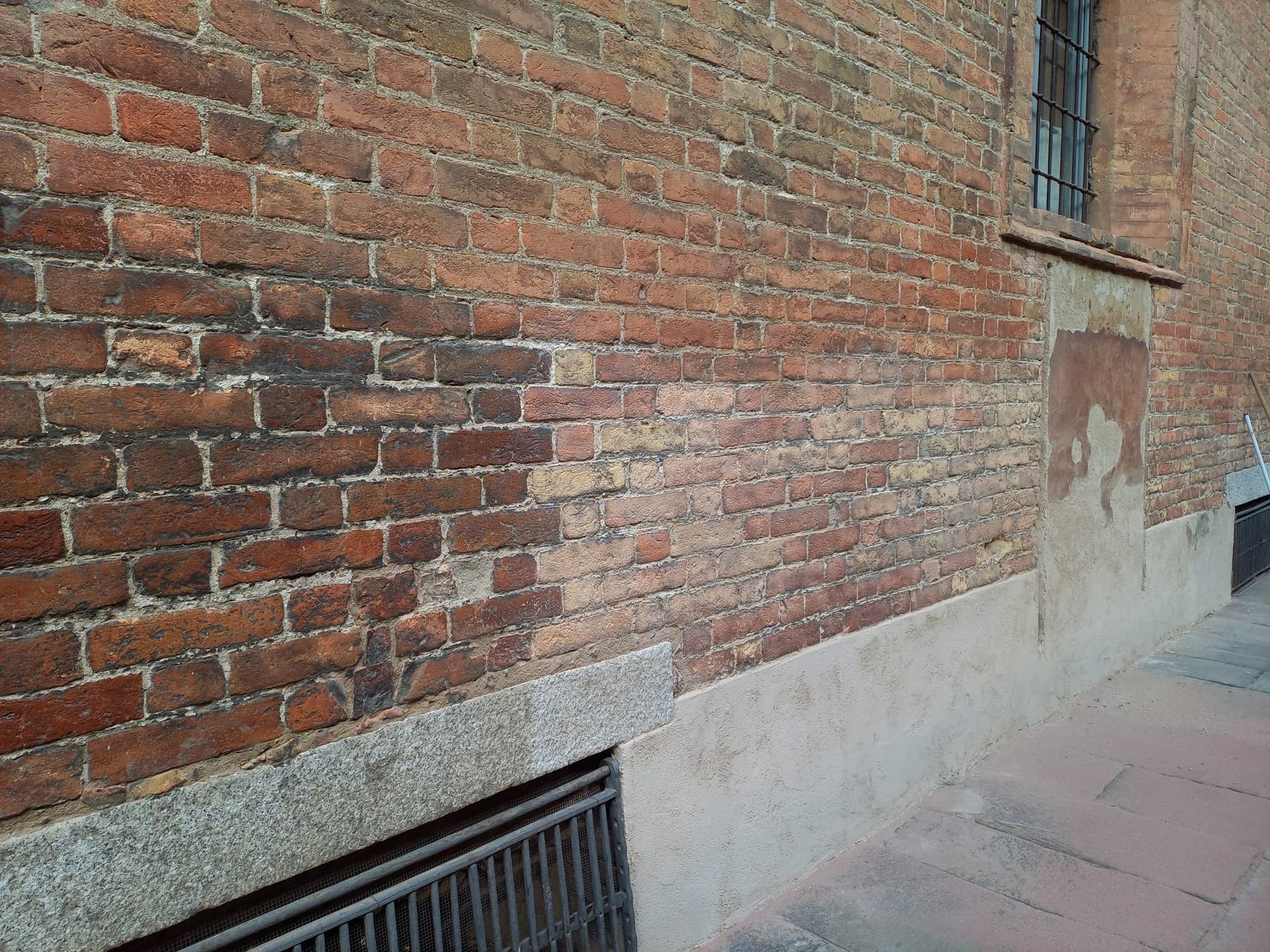 Muro di mattoni con macchie scure, finestra e base in pietra.