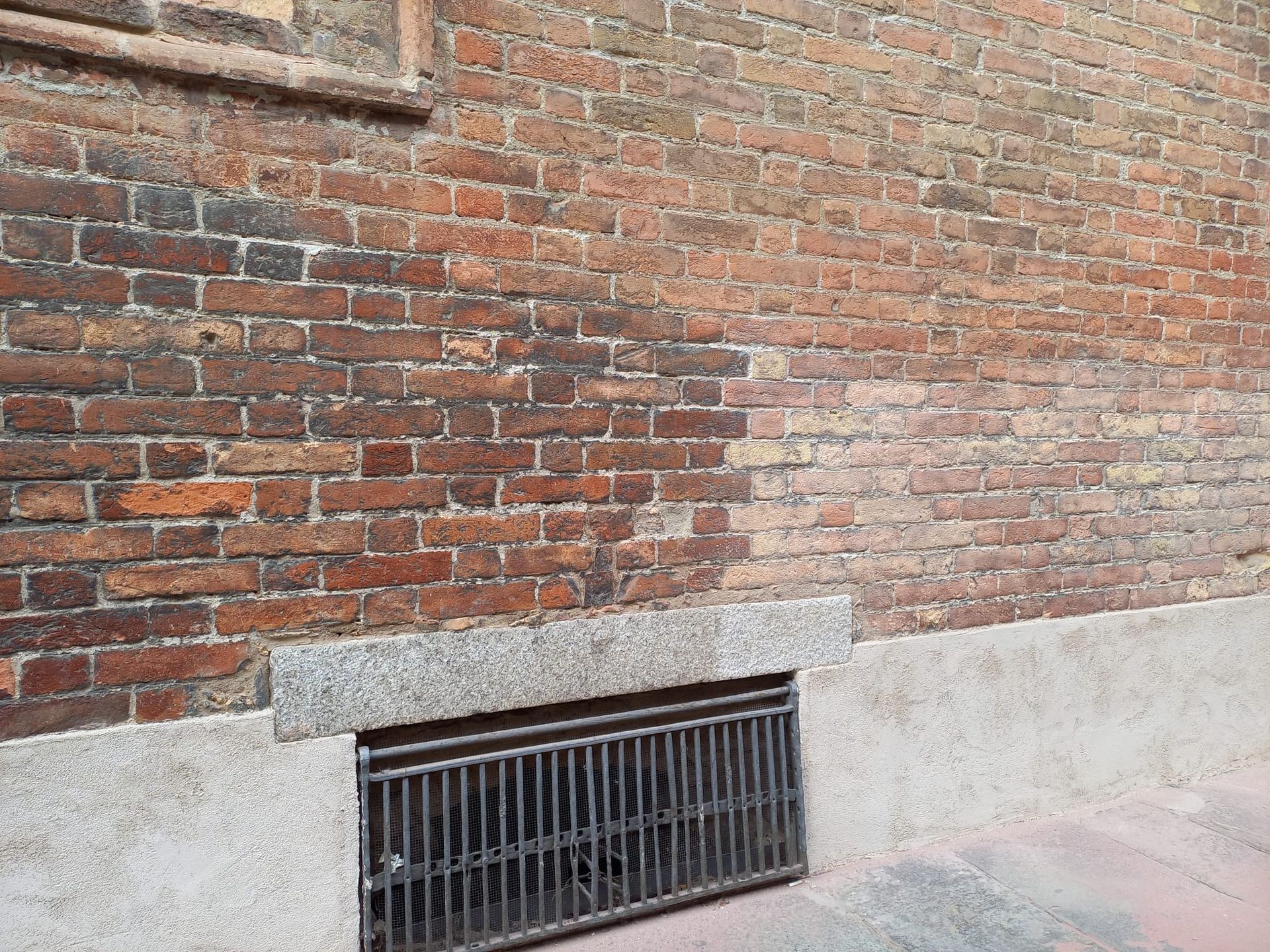 Muro di mattoni con una griglia metallica alla base, dettaglio in pietra grigia.