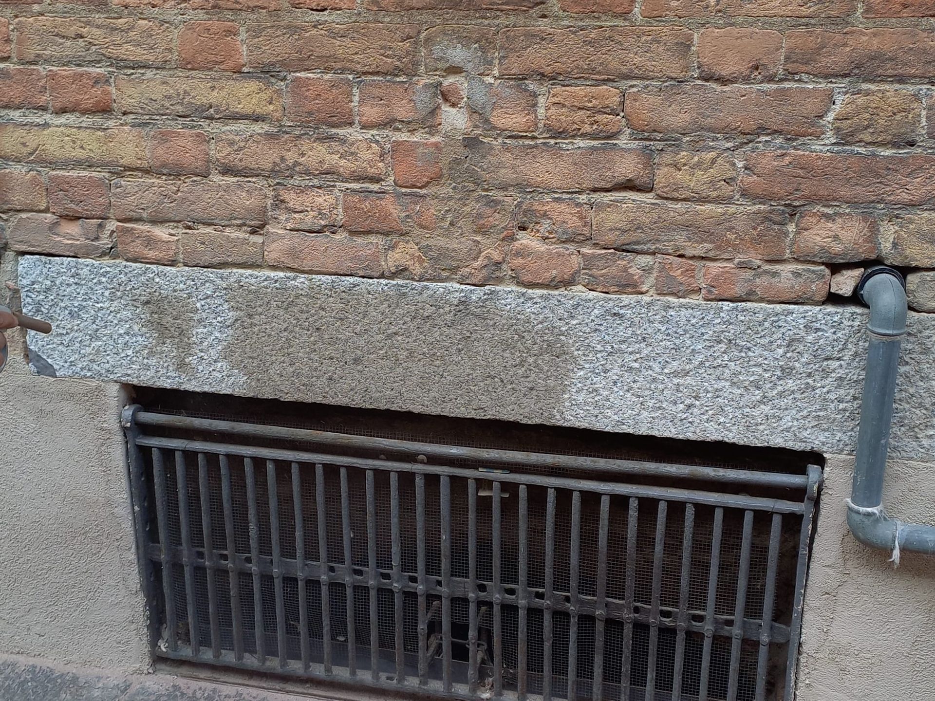 Esterno di un edificio in mattoni con una sporgenza in pietra, una grata metallica e un tubo di scarico.
