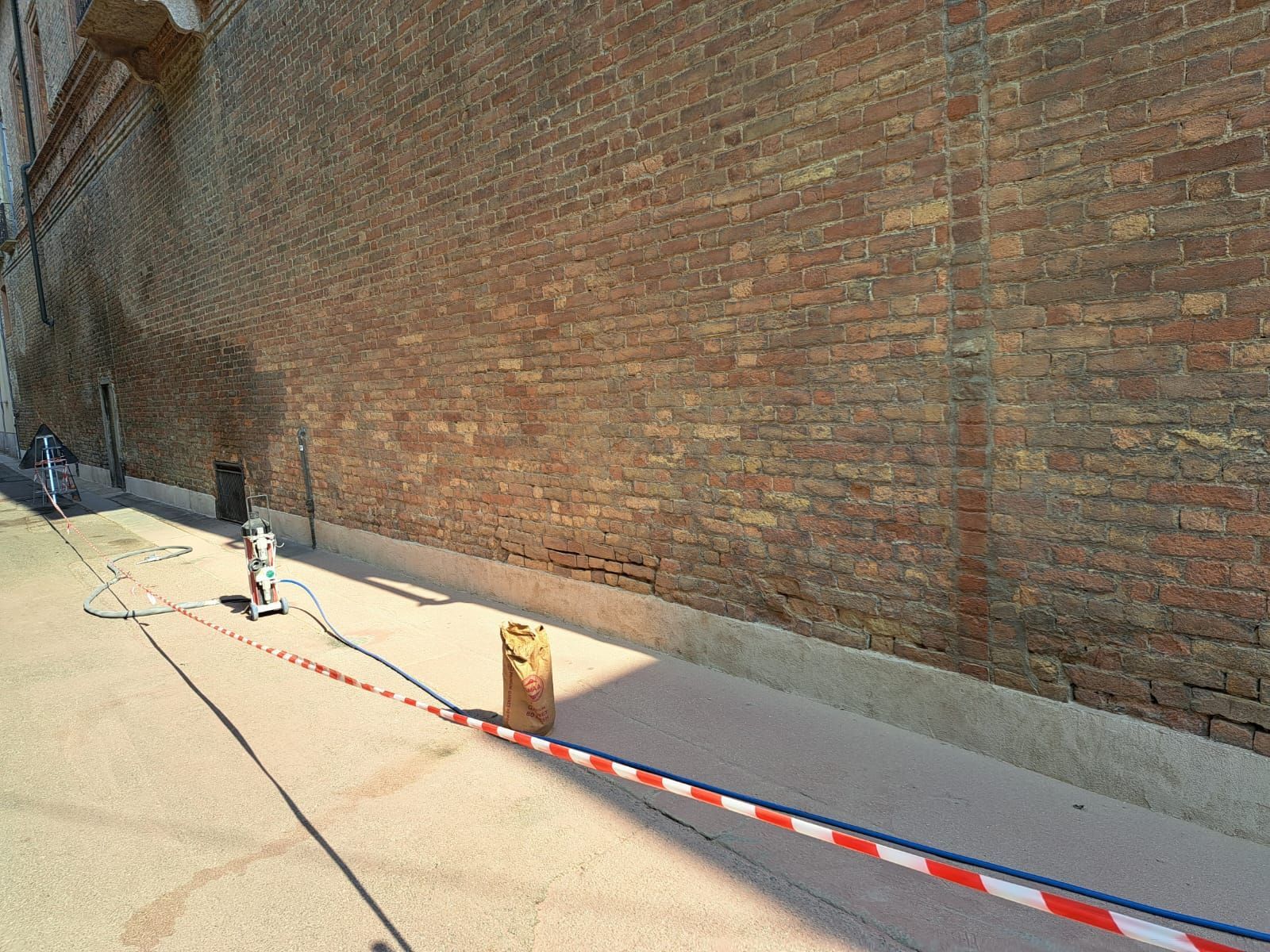 Muro di mattoni, cantiere; lungo la base del muro viene posata una base di cemento, è presente del nastro di barriera rosso/bianco.