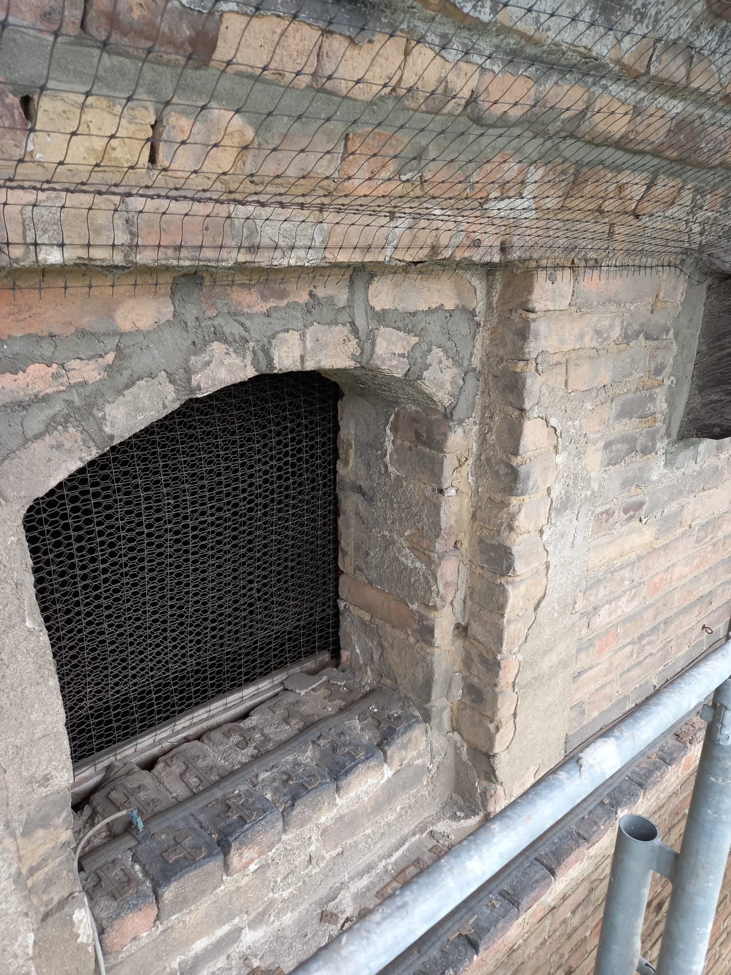 Muro di mattoni con finestra ad arco, rete metallica e impalcature.