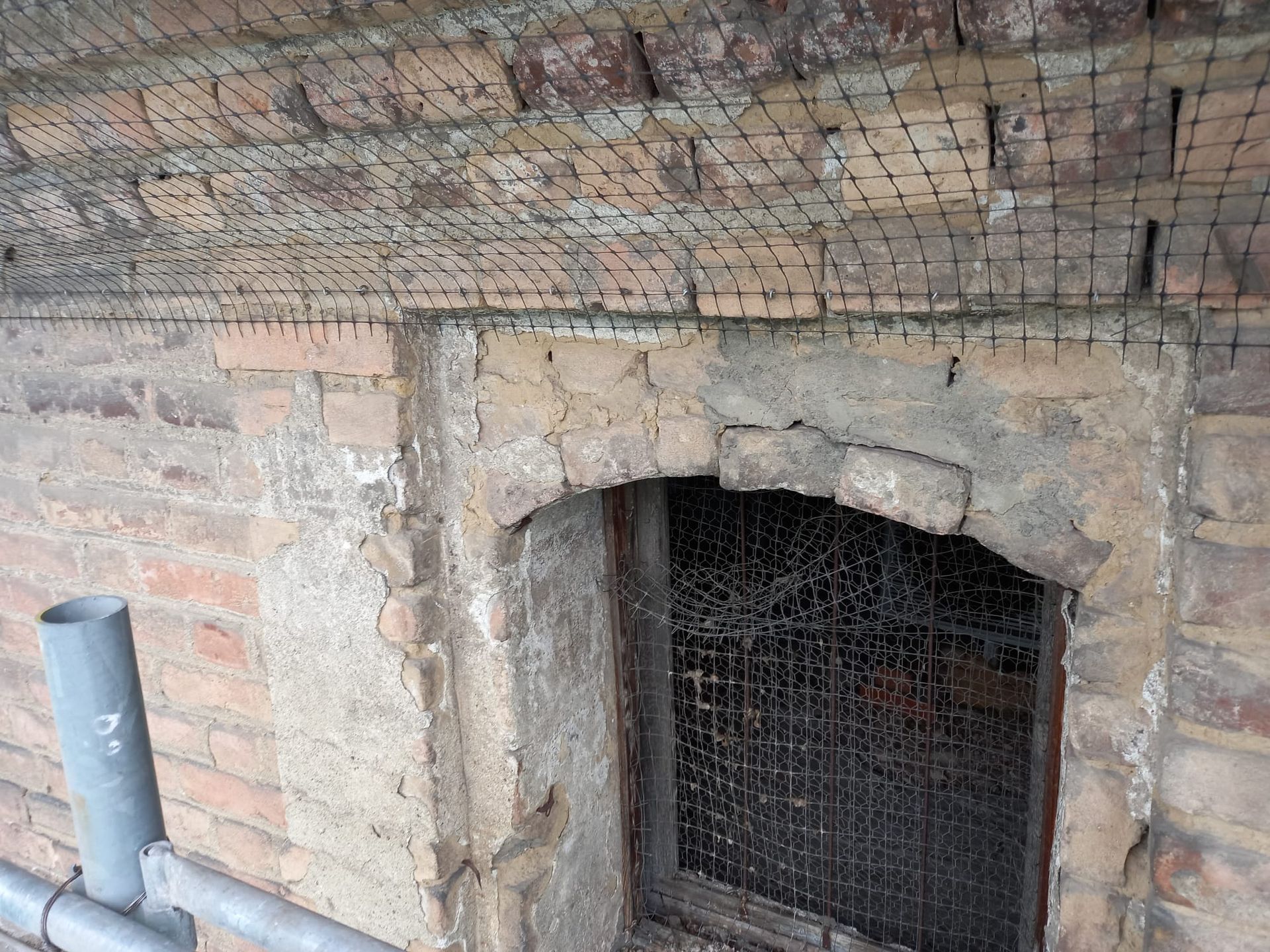 Muro di mattoni con finestra ad arco, protetto da rete metallica. Impalcature visibili sotto.