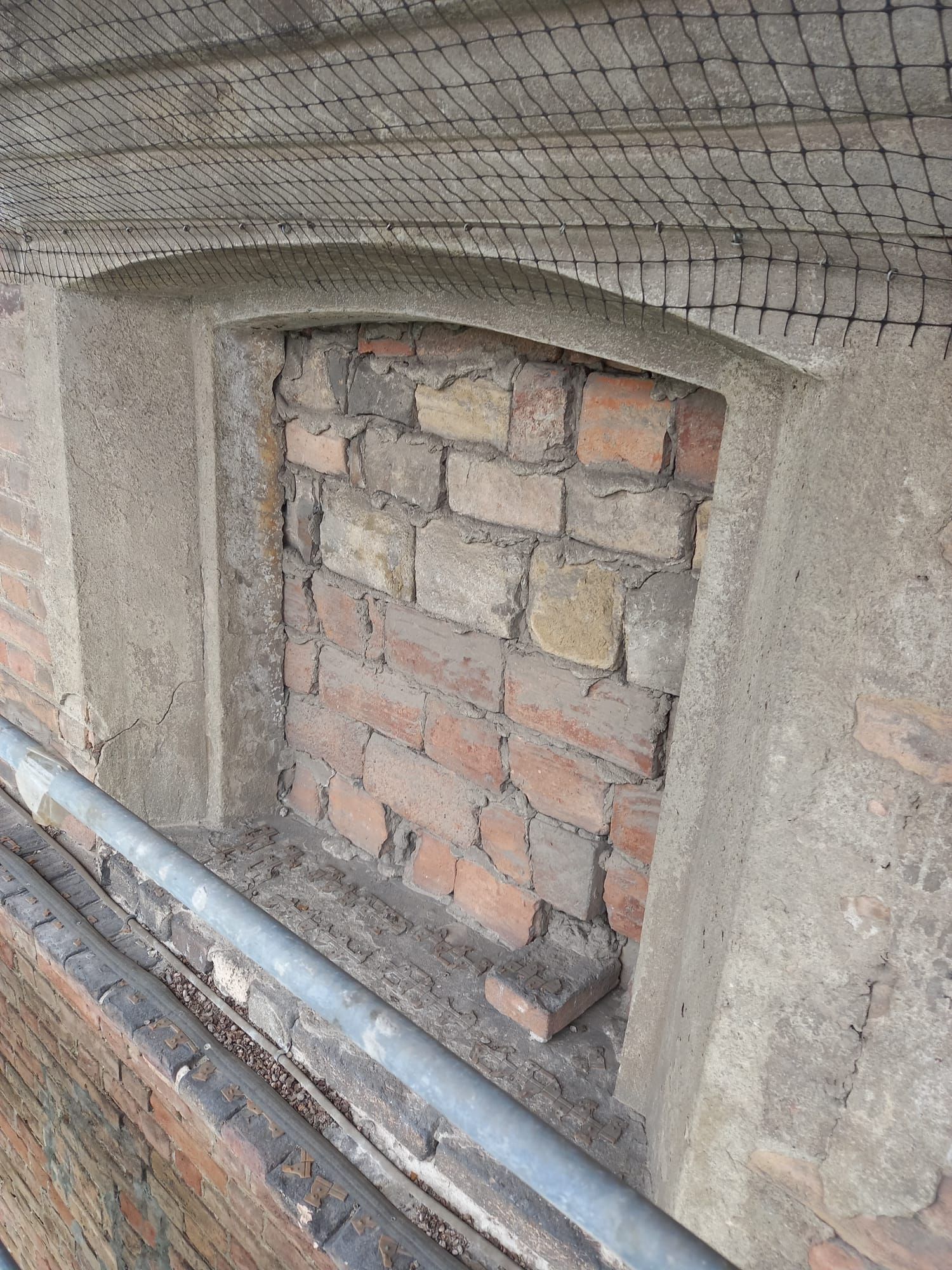 Finestra ad arco murata su un muro di mattoni. L'apertura è riempita di mattoni rossi e beige, con una cornice di cemento.