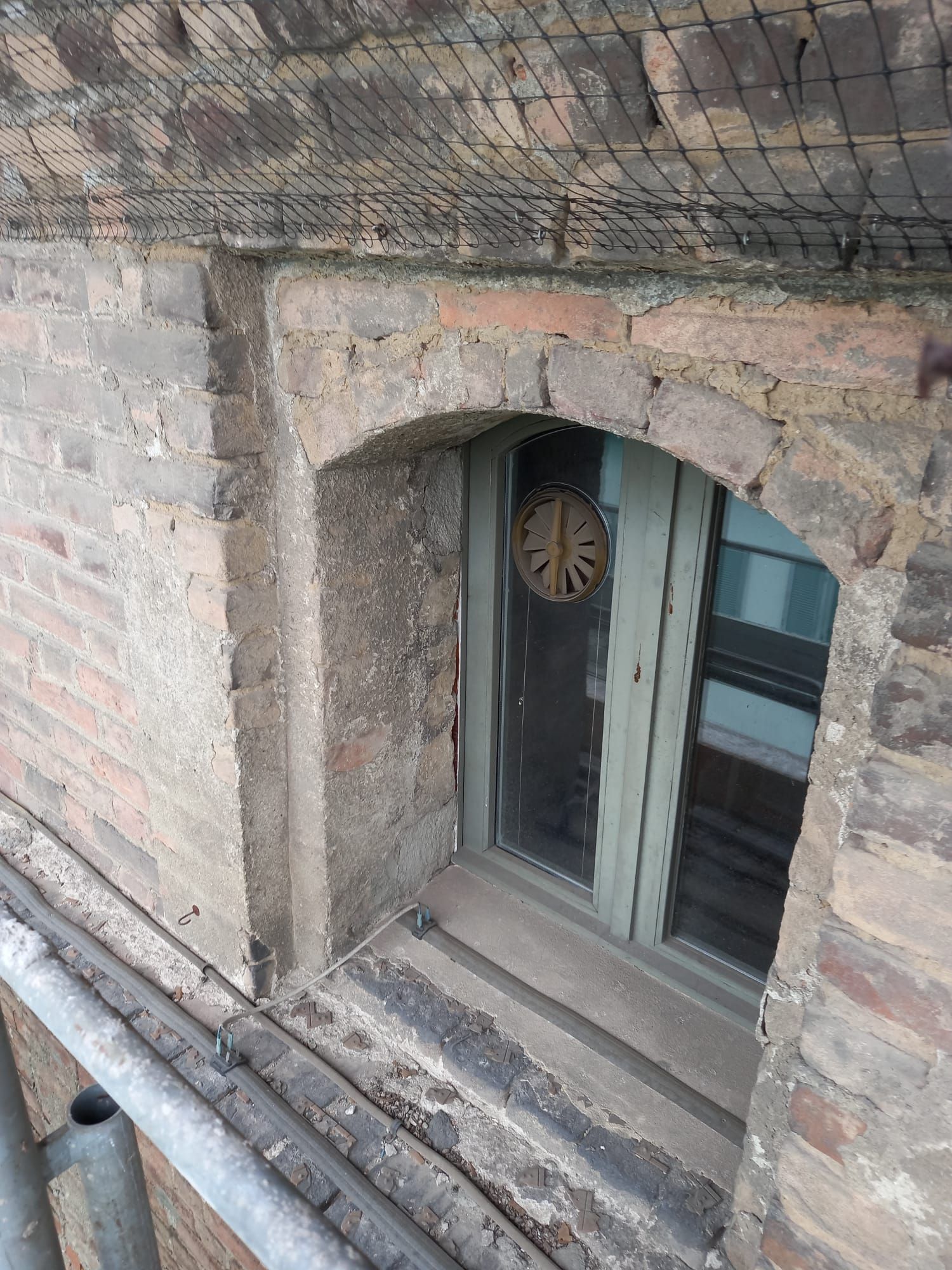 Esterno dell'edificio in mattoni con finestra ad arco, struttura in legno, sormontato da rete anti-uccelli e impalcature metalliche.
