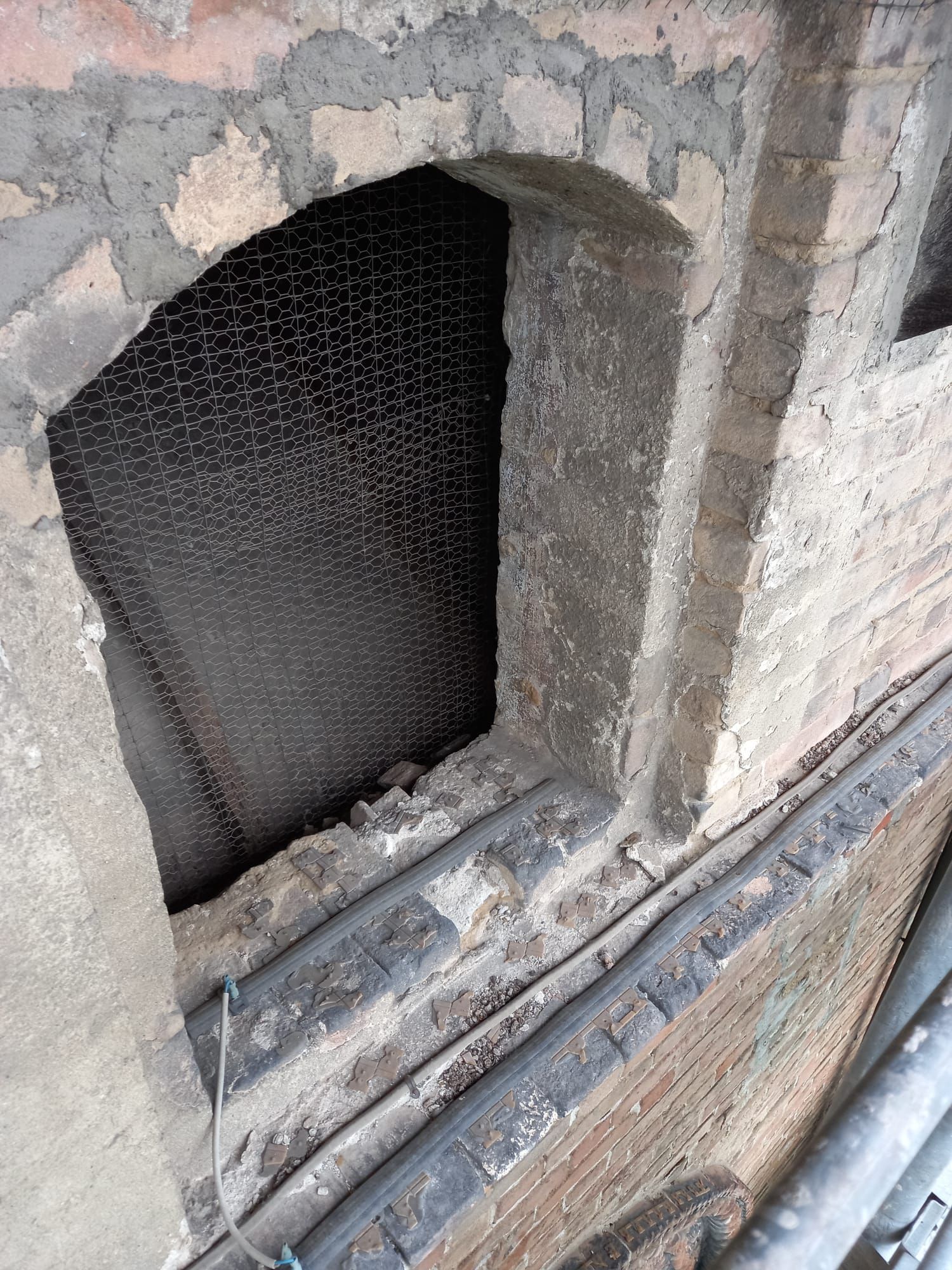 Apertura in mattoni coperta da rete scura, malta grigia e muratura visibili.