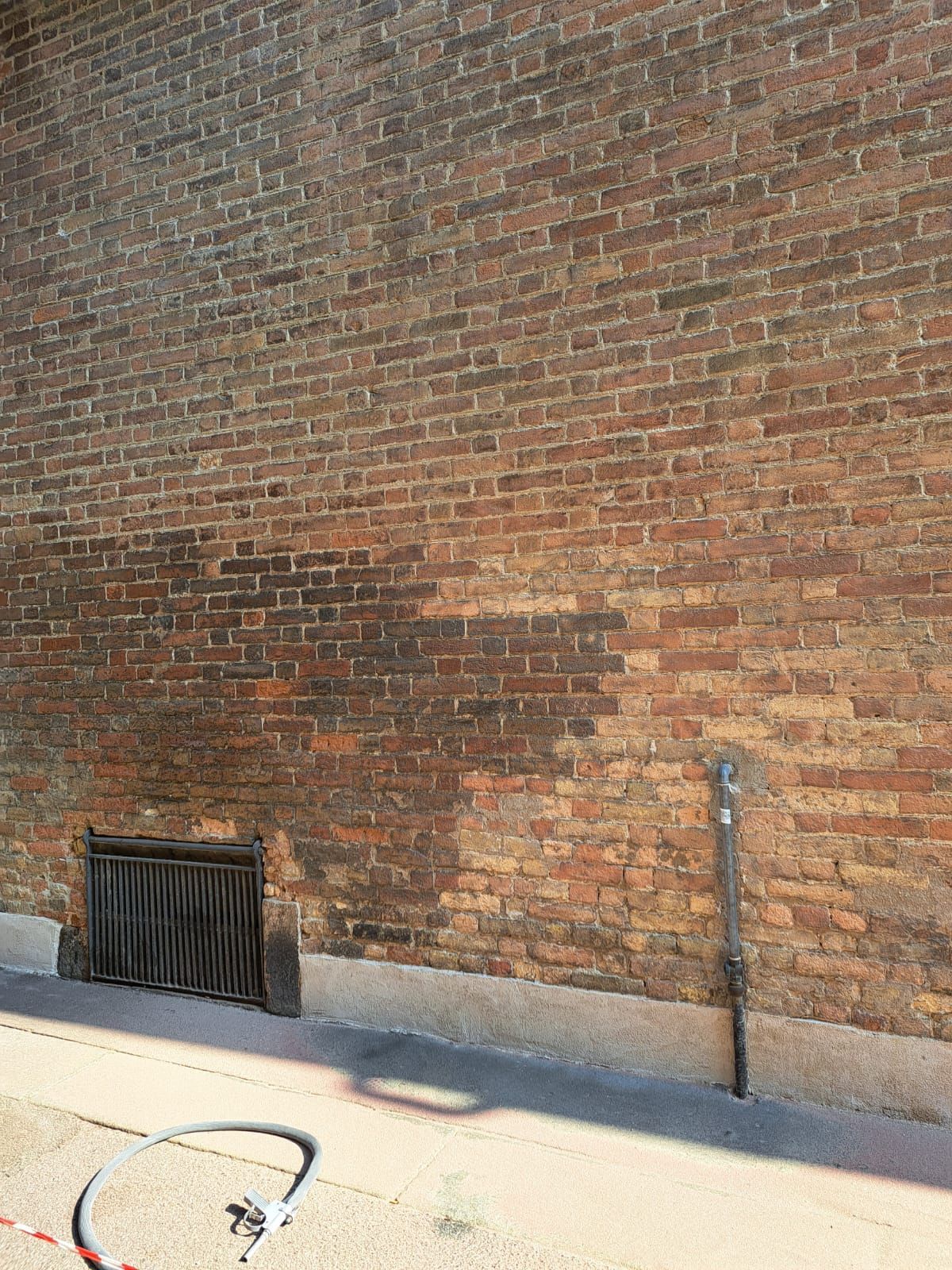 Muro di mattoni con macchia scura, una grata di metallo, un tubo flessibile e una conduttura.