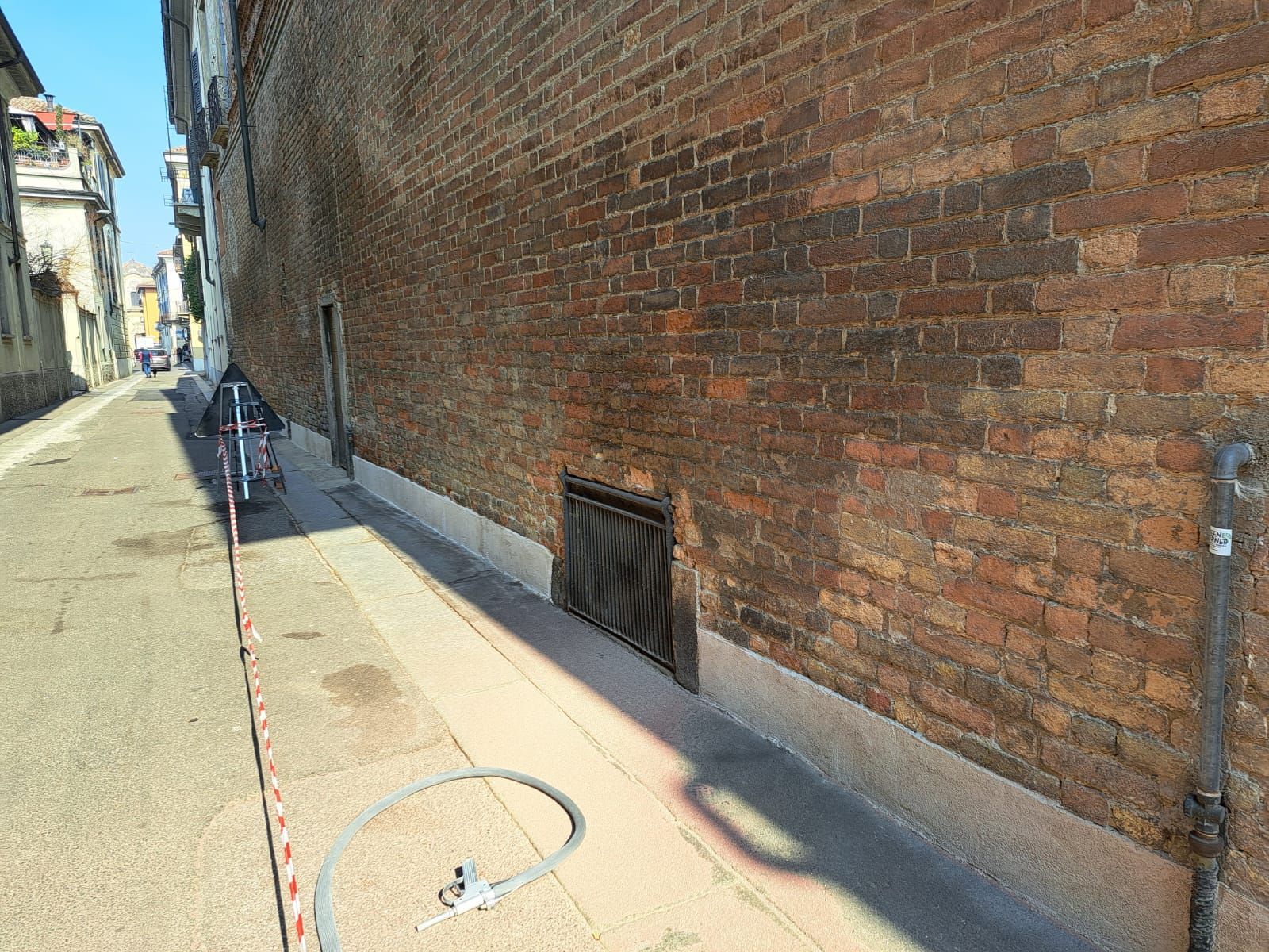 Una strada stretta con un muro di mattoni su un lato. Una bicicletta e un tubo dell'acqua. Una giornata di sole.
