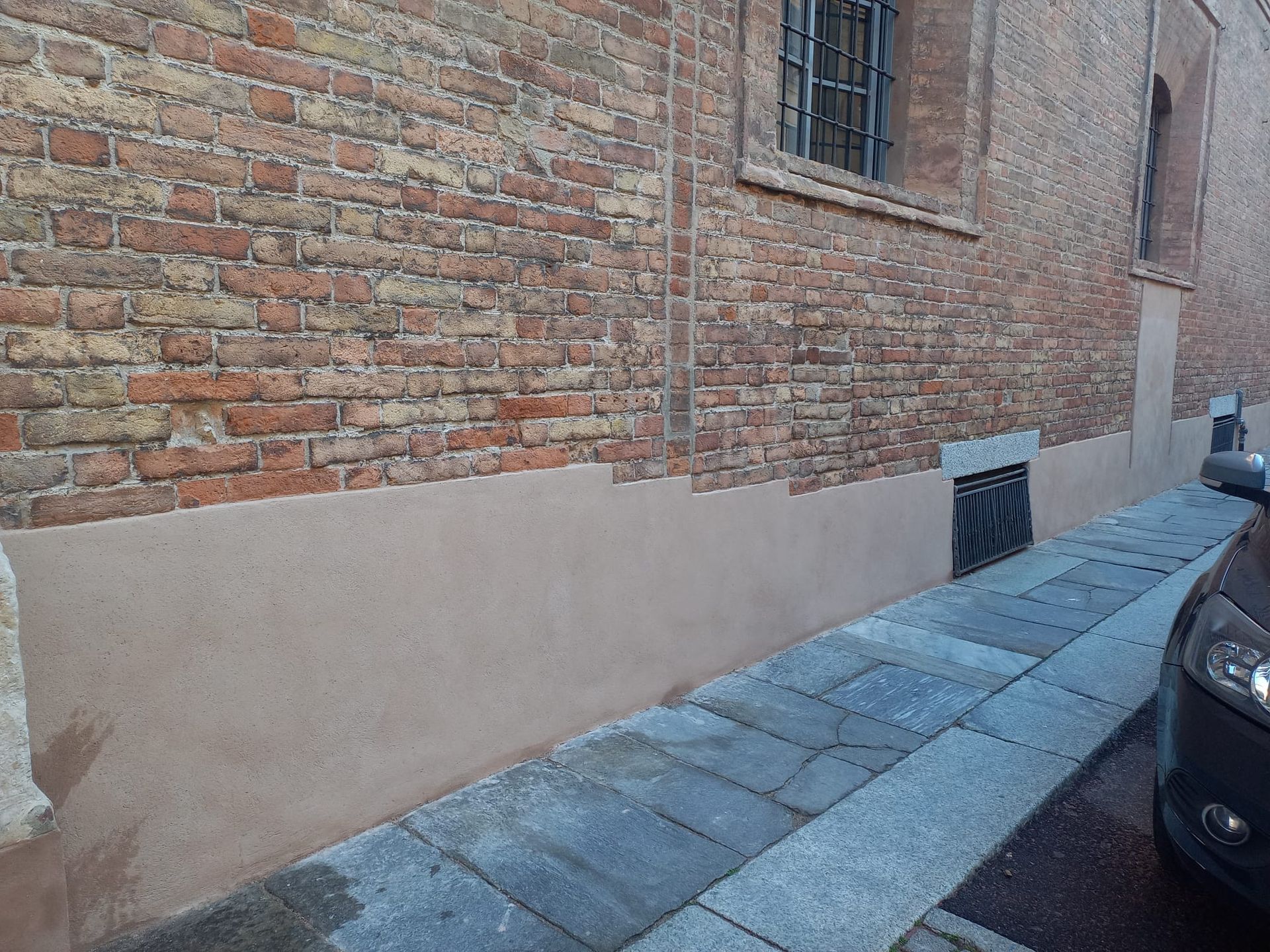 Muro di mattoni di un edificio con marciapiede, auto parcheggiata sulla destra.