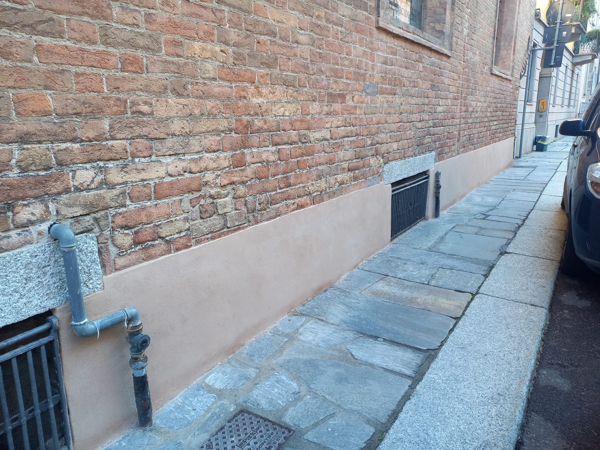 Esterno di un edificio in mattoni con marciapiede stretto. Pietra grigia, mattoni rossi, base marrone chiaro. Un'auto è parcheggiata sulla destra.