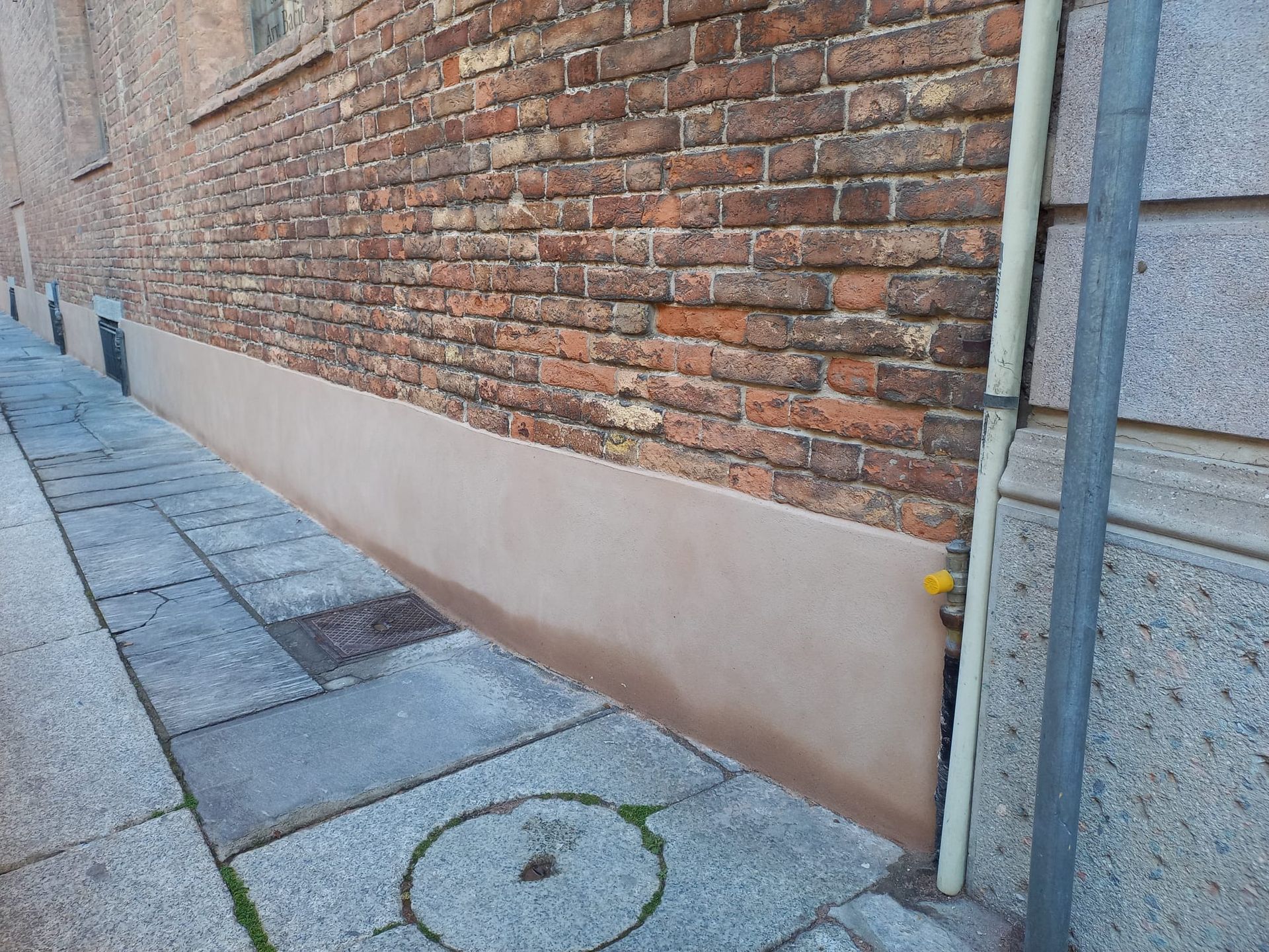 Muro di mattoni con base marrone chiaro, accanto a un marciapiede pavimentato e a un tubo verticale.
