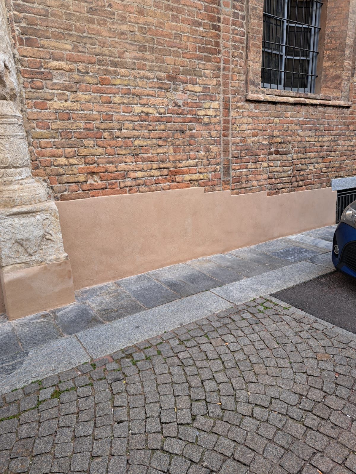 Muro di mattoni con la parte inferiore color marrone chiaro, strada acciottolata e un'auto parzialmente visibile.