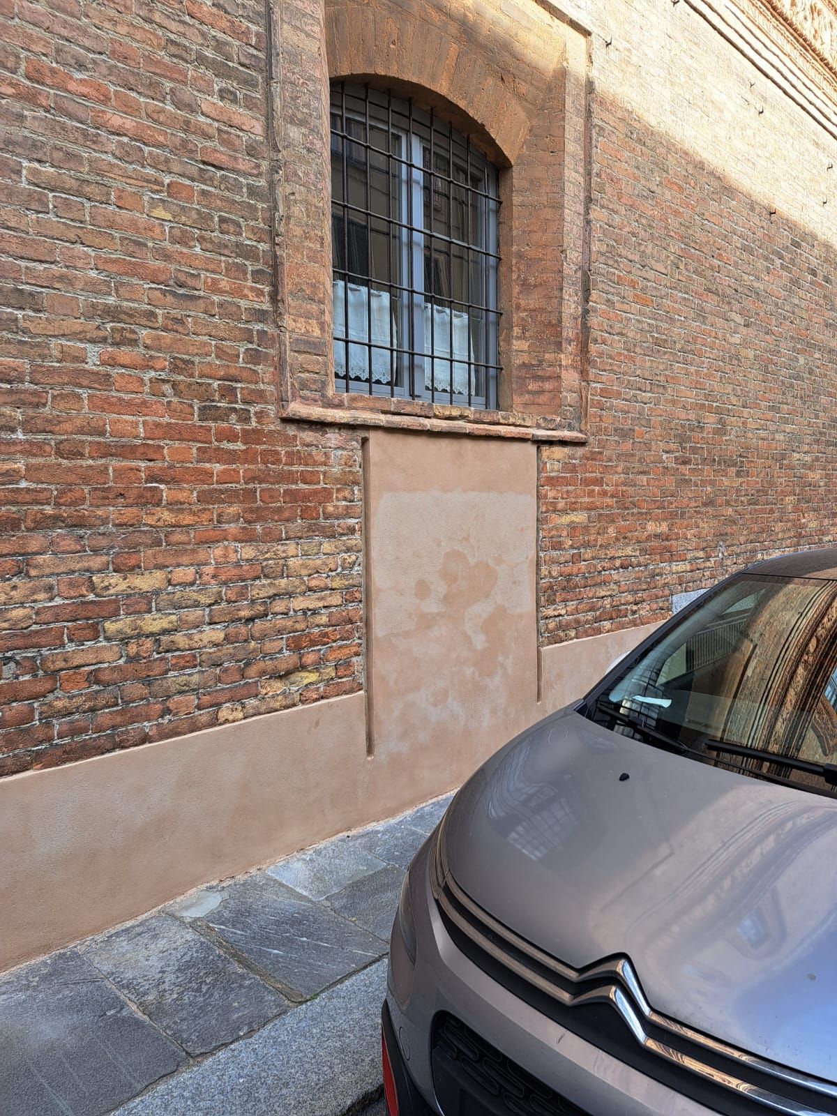 Edificio in mattoni rossi con una finestra sbarrata e una macchia marrone chiaro dove un tempo c'era una porta; auto parcheggiata di fronte.