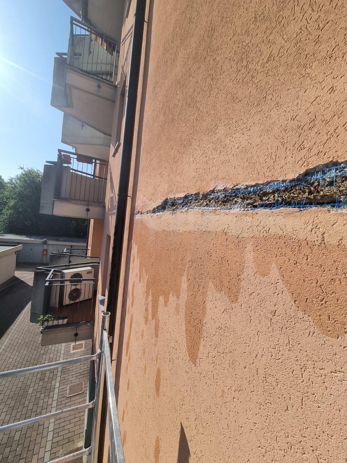 Muro esterno con una giuntura scura e danneggiata, macchie d'acqua che colavano lungo l'intonaco. Sono visibili balconi e una ringhiera.