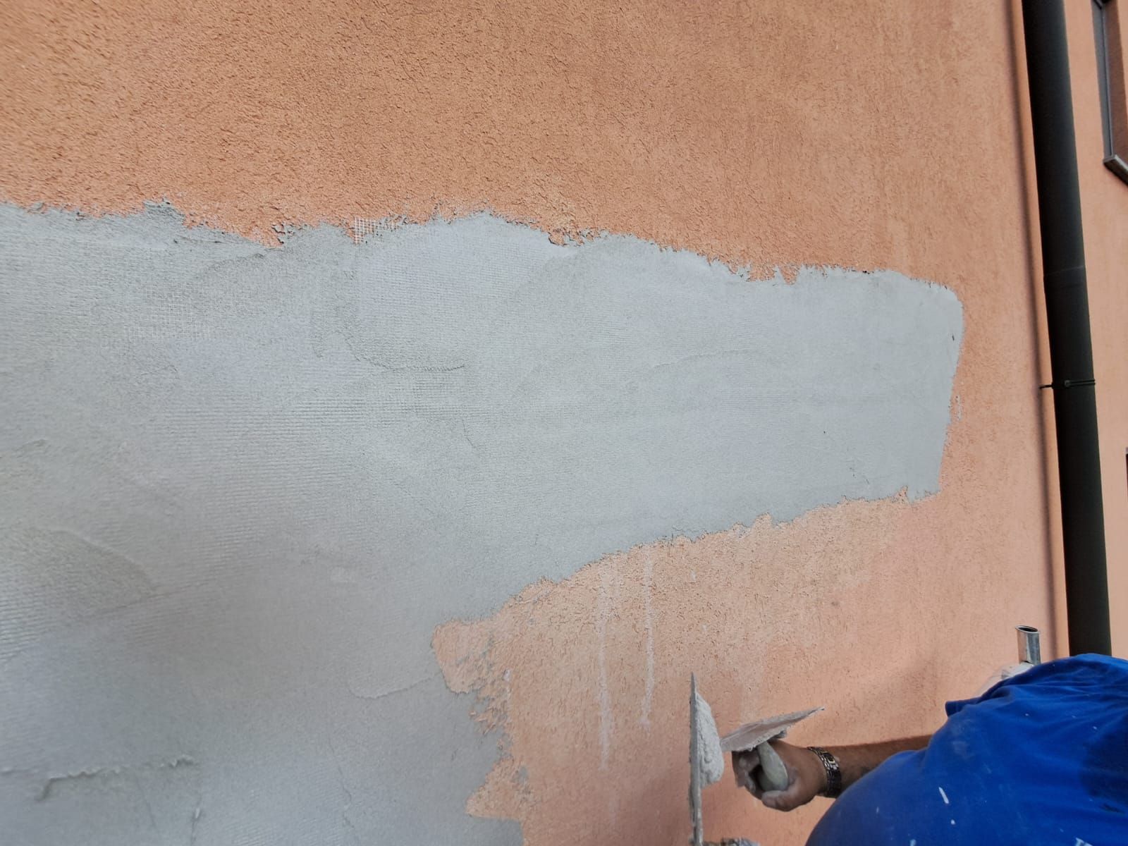 Una persona che applica con una cazzuola uno stucco grigio su una parete esterna color salmone.