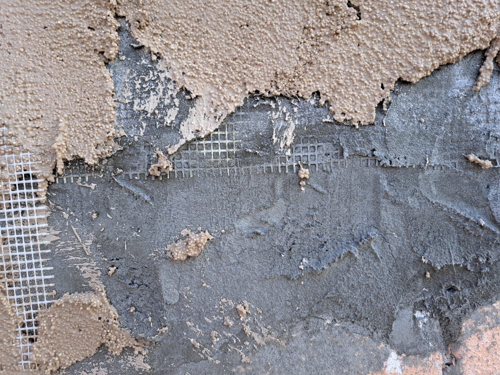 Parete in stucco danneggiata con strato di rinforzo in rete a vista e intonaco sgretolato.