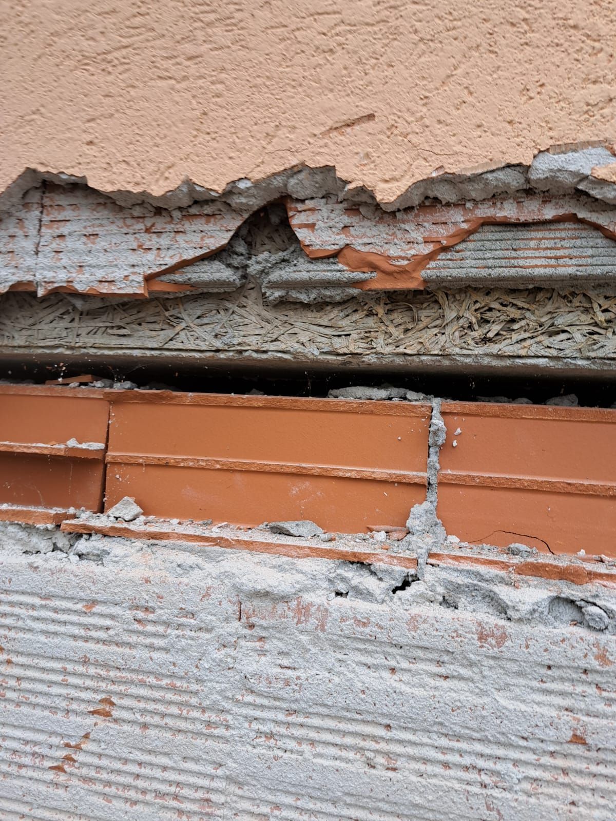 Muro esterno danneggiato: intonaco arancione sgretolato su mattoni a vista, con una fessura visibile e cemento grigio usurato.