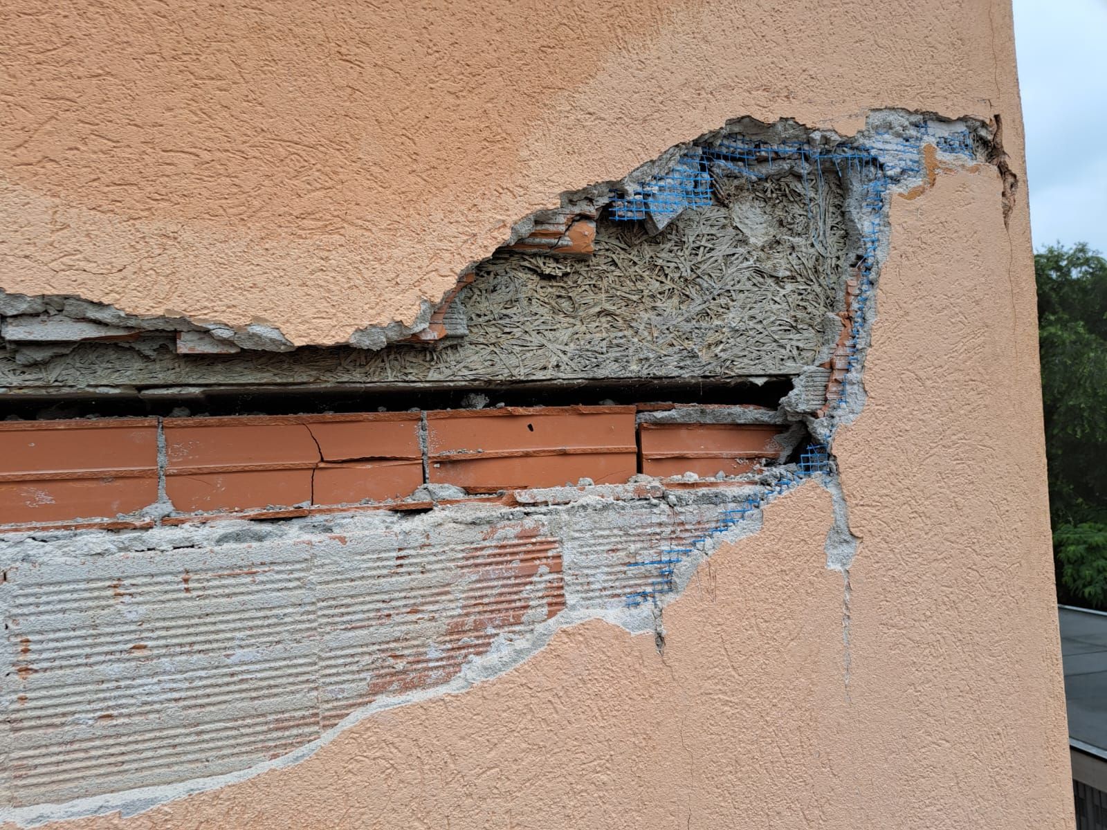 Muro esterno danneggiato che lascia a vista mattoni e malta. Stucco color pesca e contorni blu contrastano con i mattoni rossi.