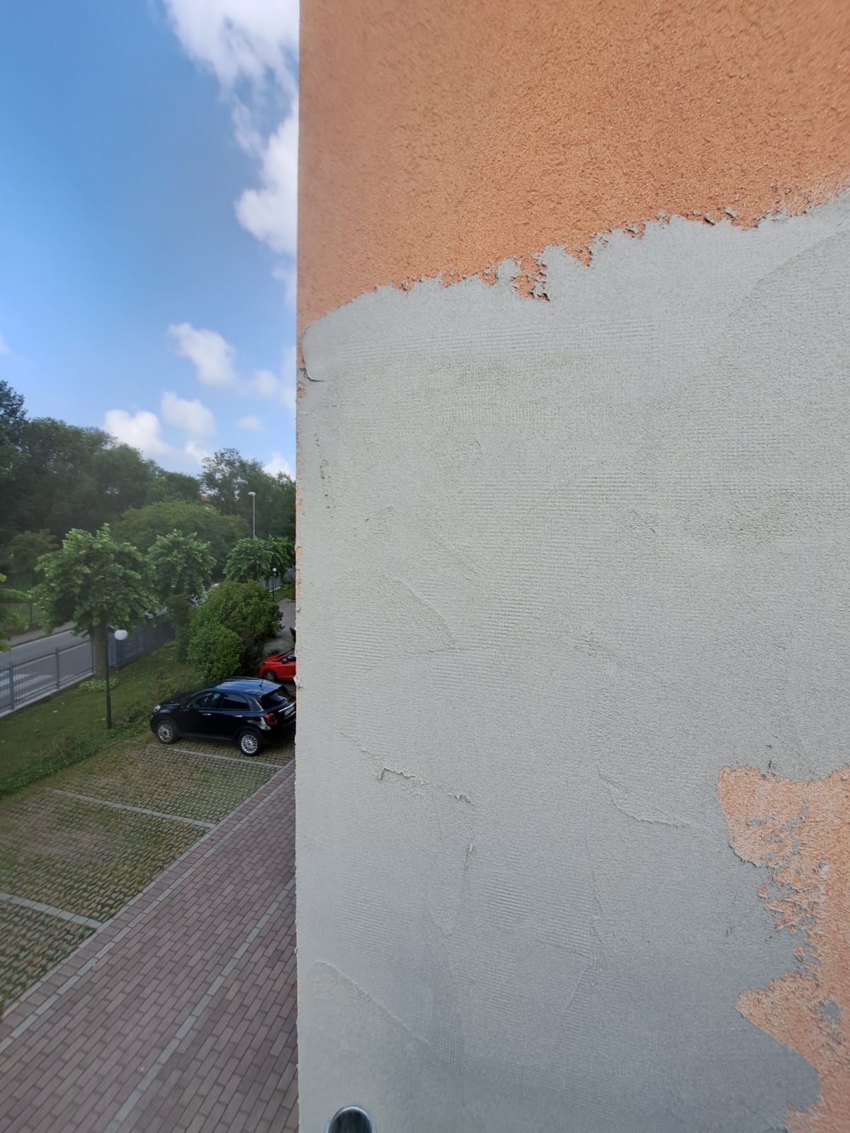 La vista da un edificio mostra un parcheggio con auto e una fila di alberi sullo sfondo di un cielo azzurro nuvoloso.