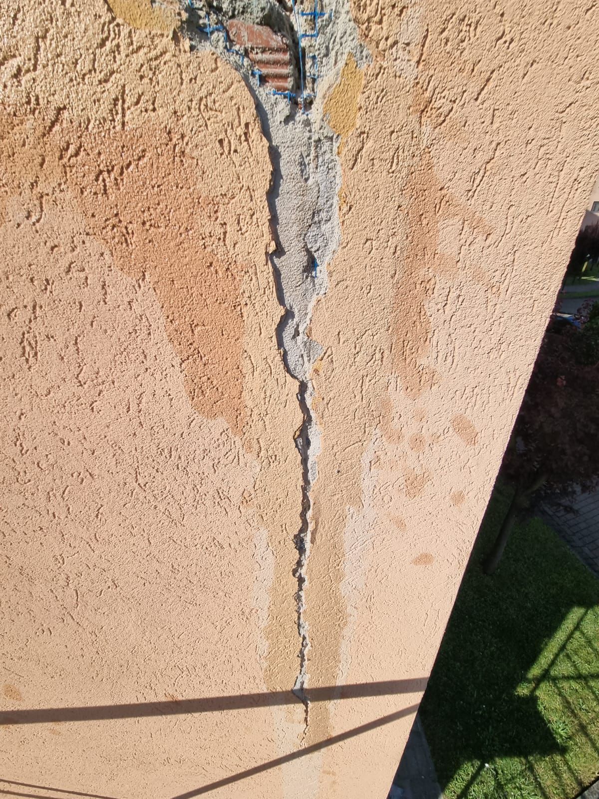 Crepa verticale nel muro esterno in stucco, di colore arancione chiaro. Mostra cemento a vista con pezzi di mattoni nella parte superiore.