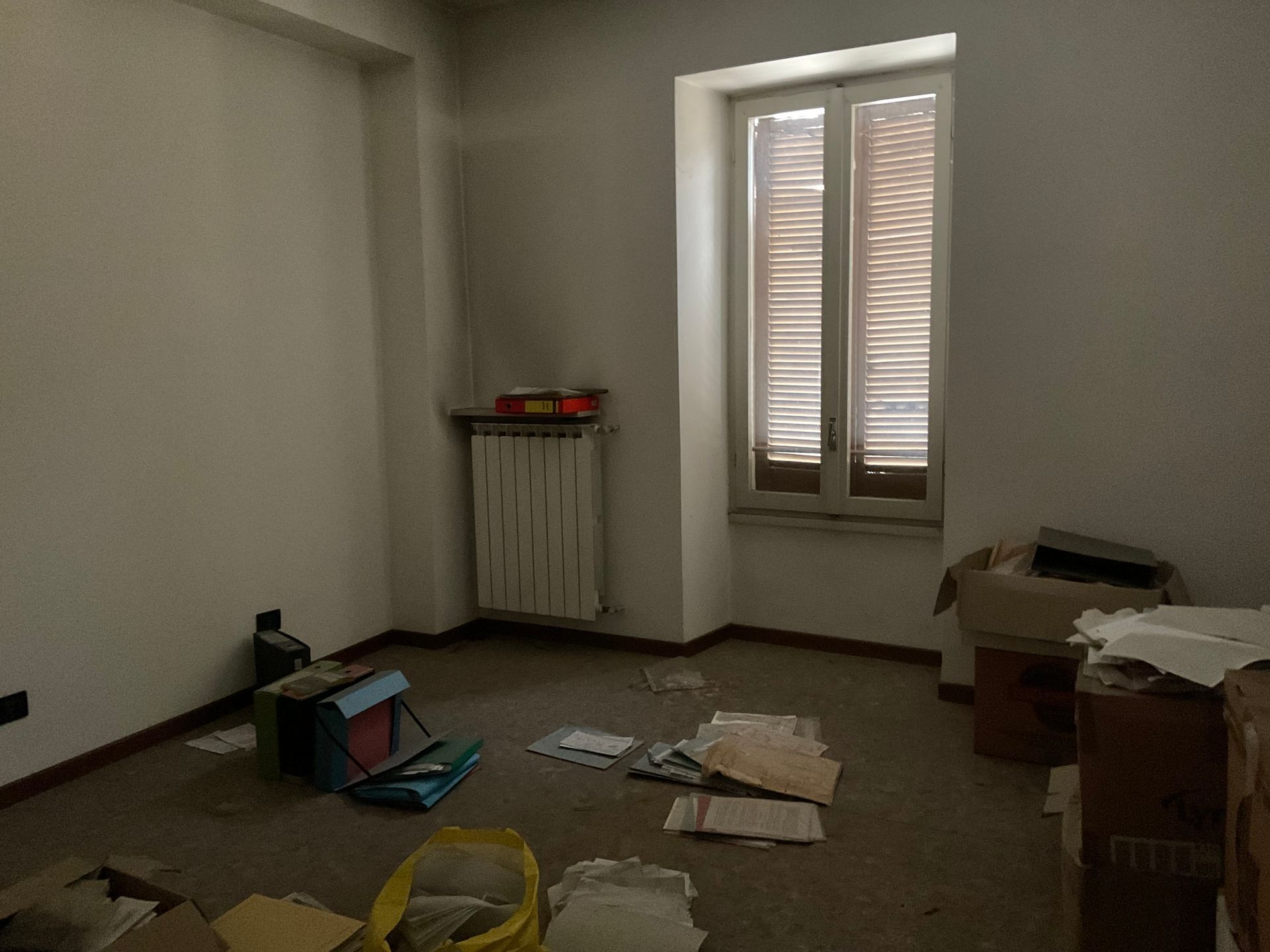 Stanza vuota con finestra, termosifone e detriti sparsi sul pavimento; probabilmente in fase di ristrutturazione.