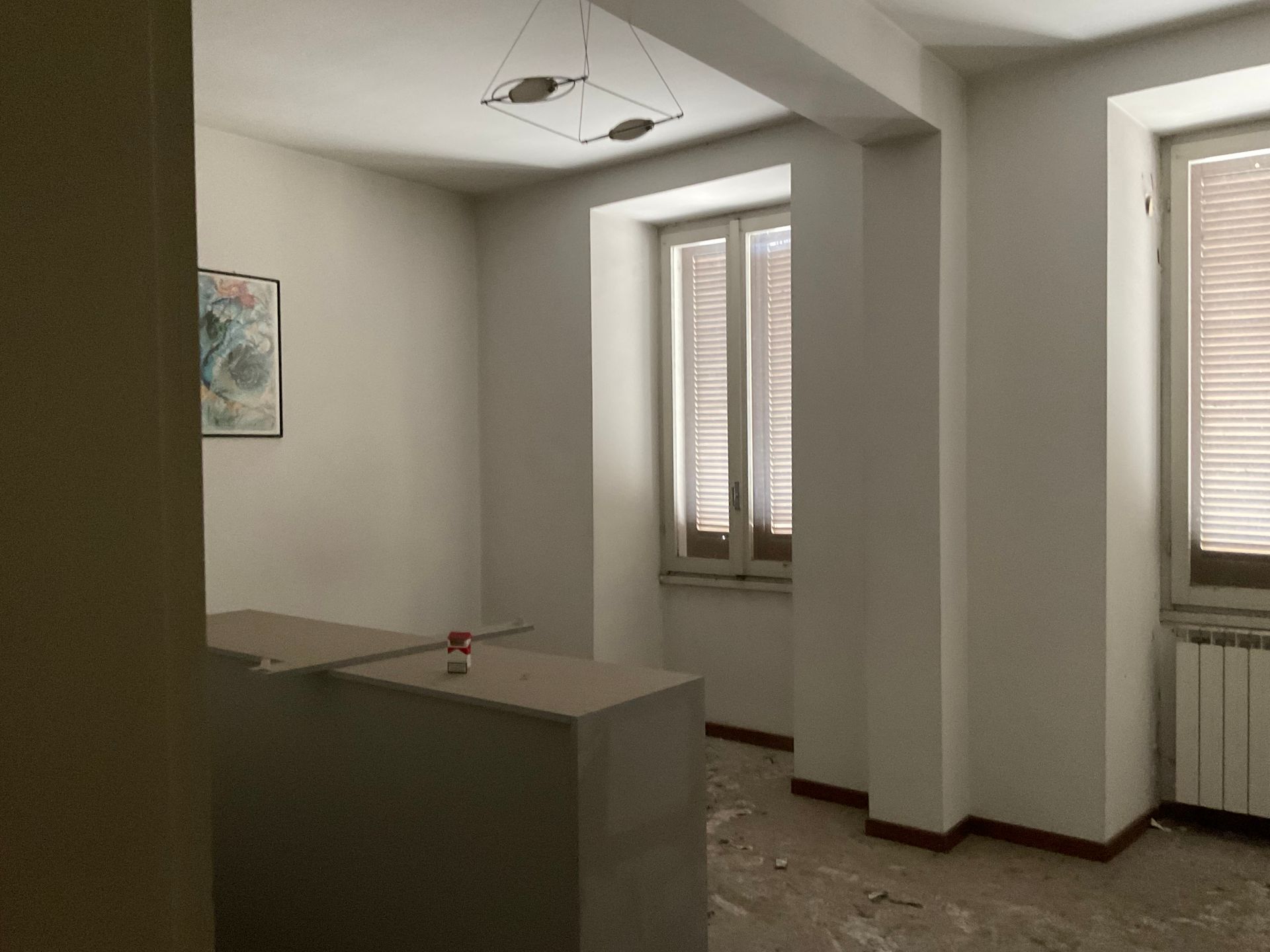 Stanza vuota con due finestre, un bancone parziale e un piccolo quadro sulla parete.