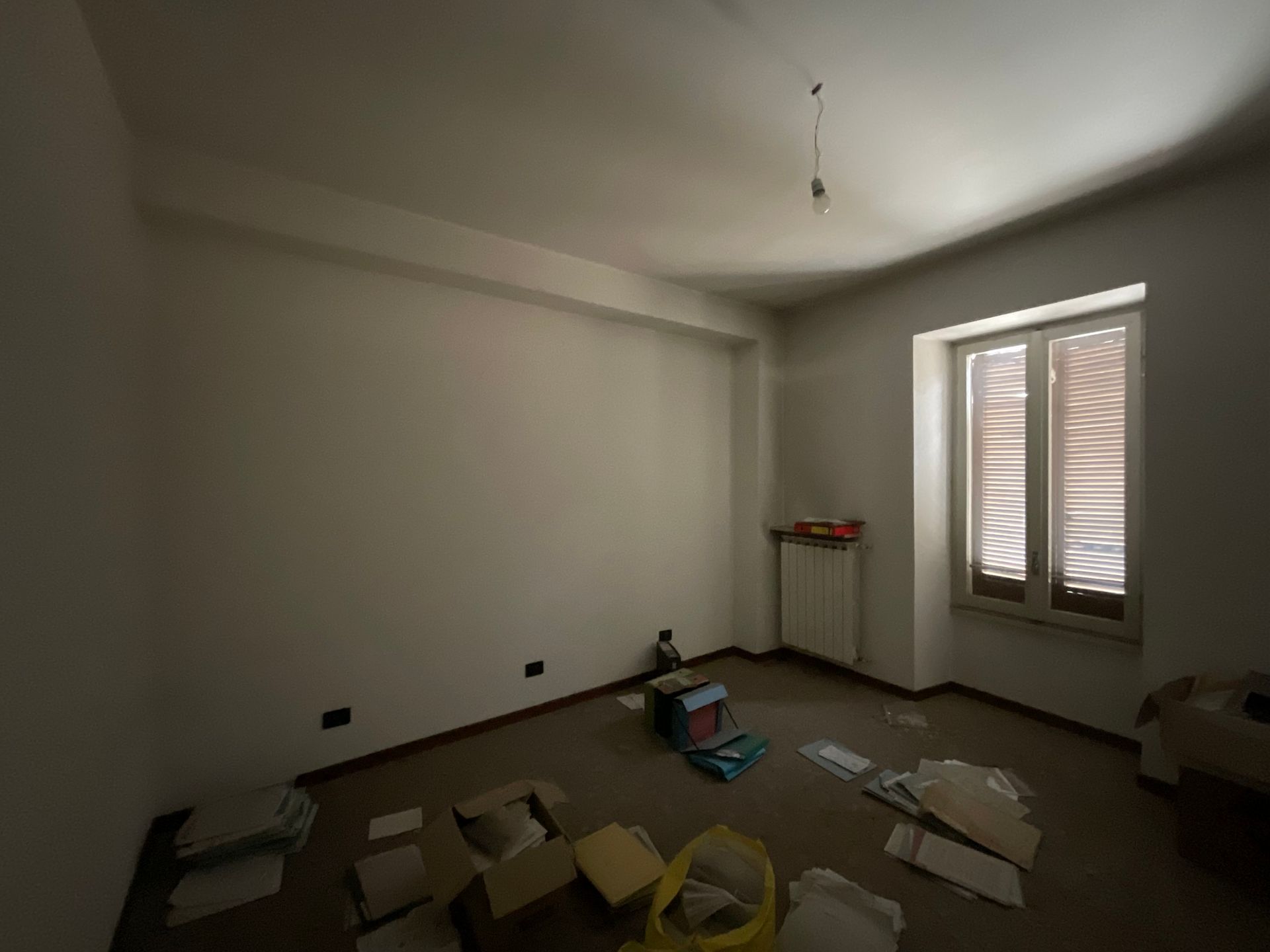 Stanza vuota con pavimento in disordine, una finestra e una sola lampadina appesa.