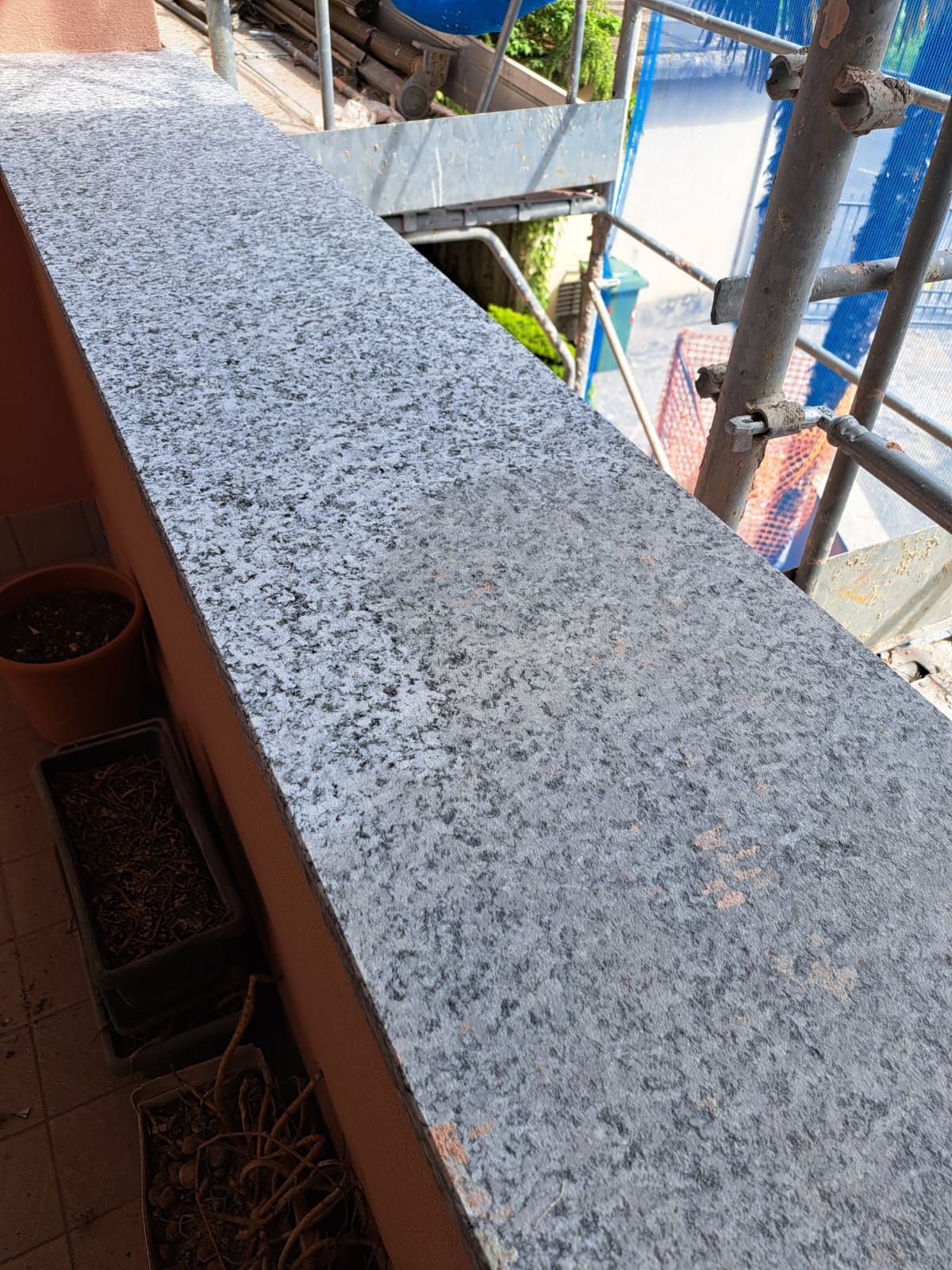 Ringhiera del balcone in granito con superficie grigia screziata, vista dall'alto. Sono visibili un'impalcatura e una pianta in vaso.