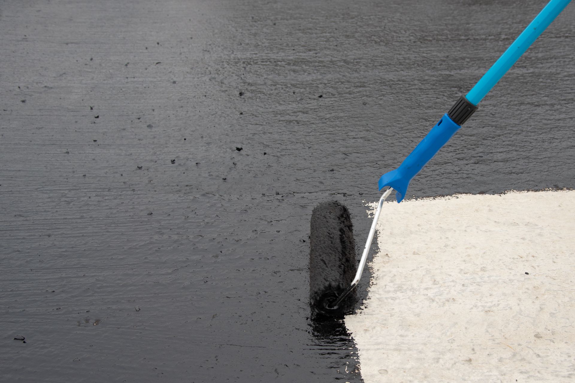 Black sealant being applied to a concrete surface with a roller and long blue handle.