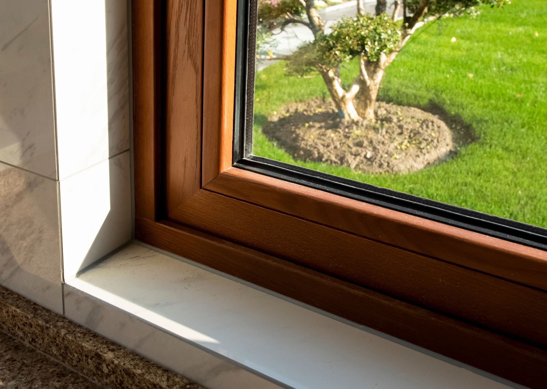 Brown window frame with a view of a green lawn and a tree. Sunlight illuminates the white window sill.
