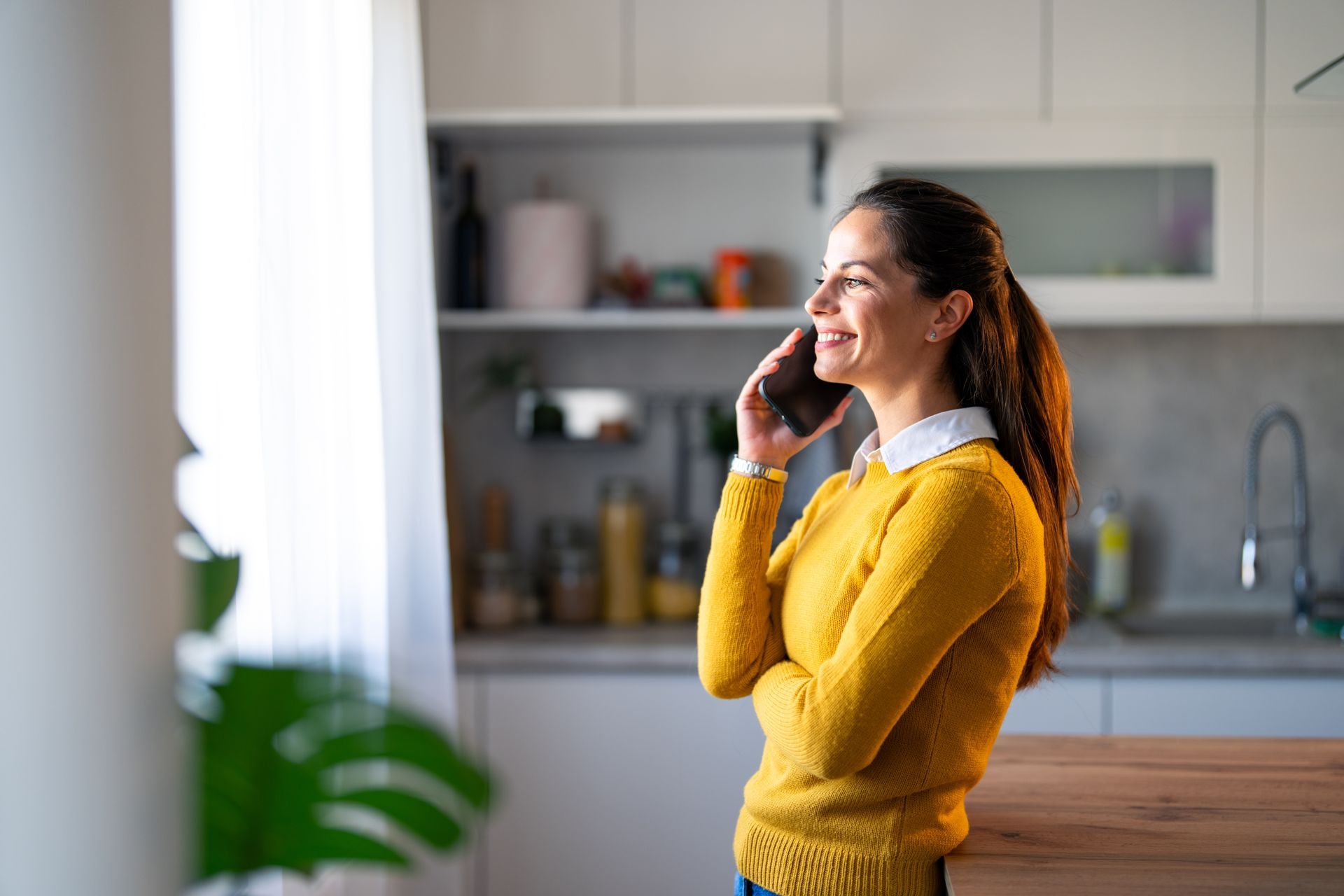 Woman in yellow sweater smiles while talking on the phone in a kitchen.