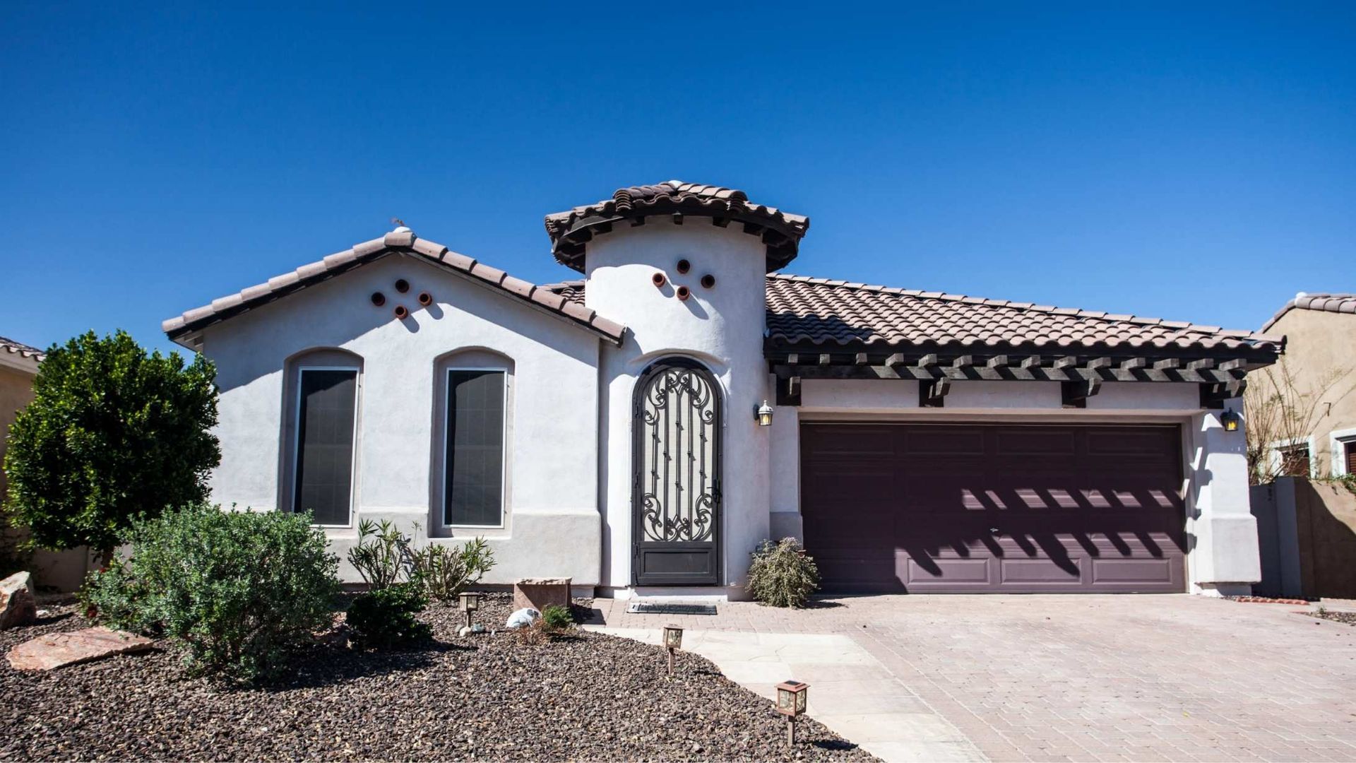 White stucco house with brown roof and garage, arched ornate front door, clear blue sky.