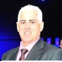 Man with white hair smiles, wearing suit and tie; dark background.