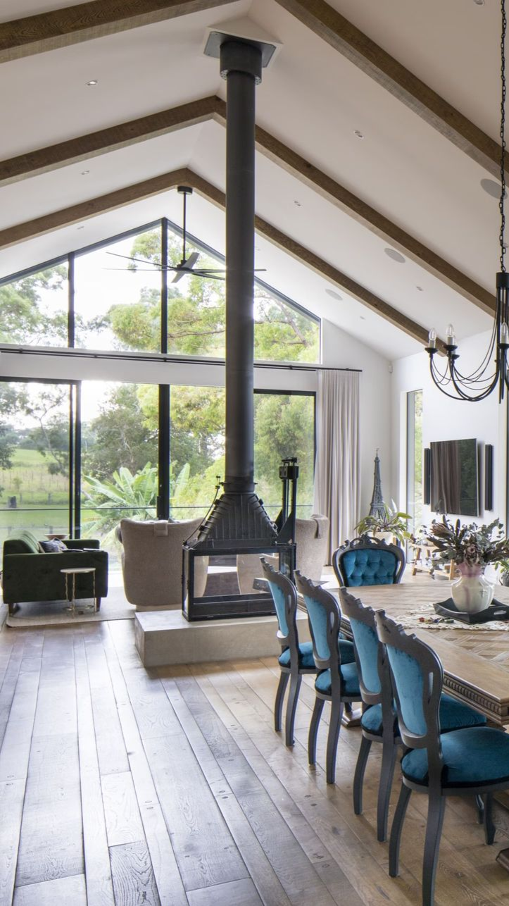 Interior of a house with a tall fireplace, vaulted ceiling, large windows, dining table with blue chairs.