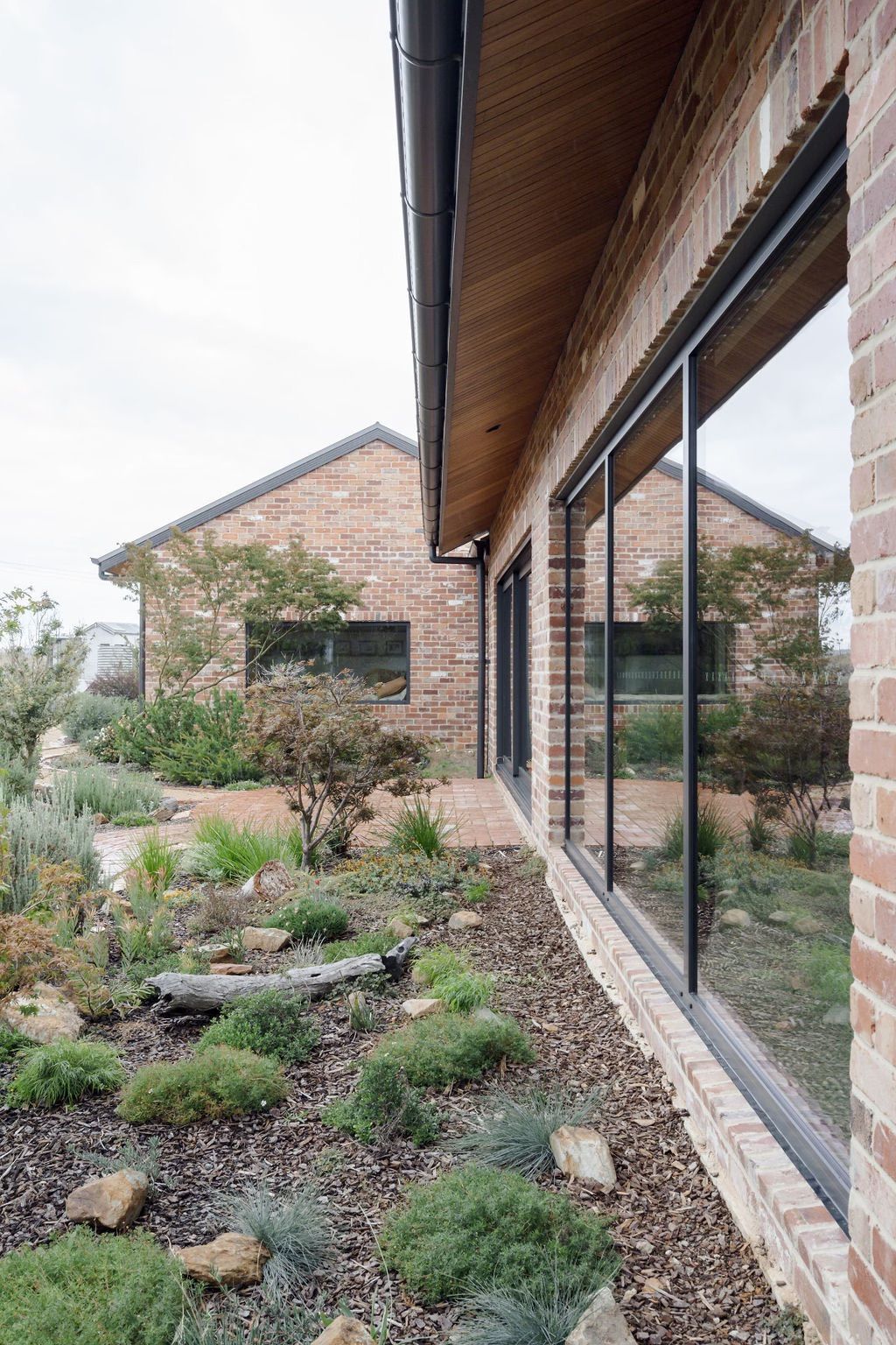 Brick building with large windows reflecting garden. Brown roof, green plants, and overcast sky.