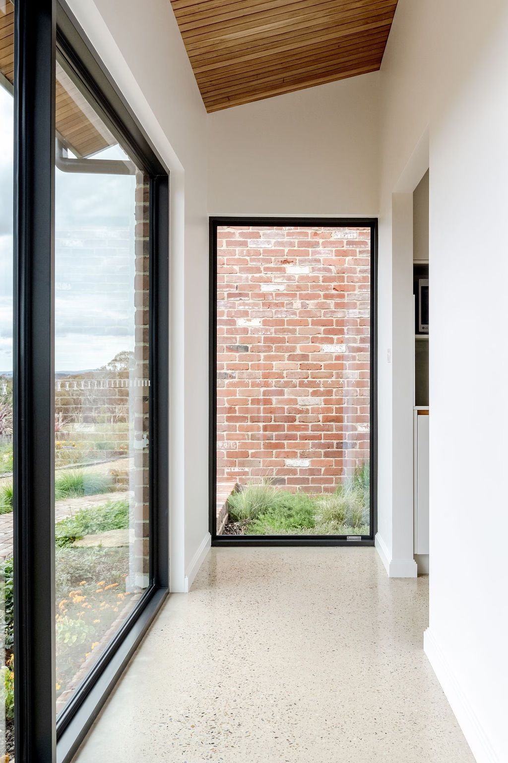 Hallway with large windows, brick wall view, concrete floor, light wood ceiling.