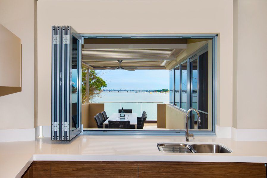 Kitchen with open folding window overlooking a patio with a table and view of water.