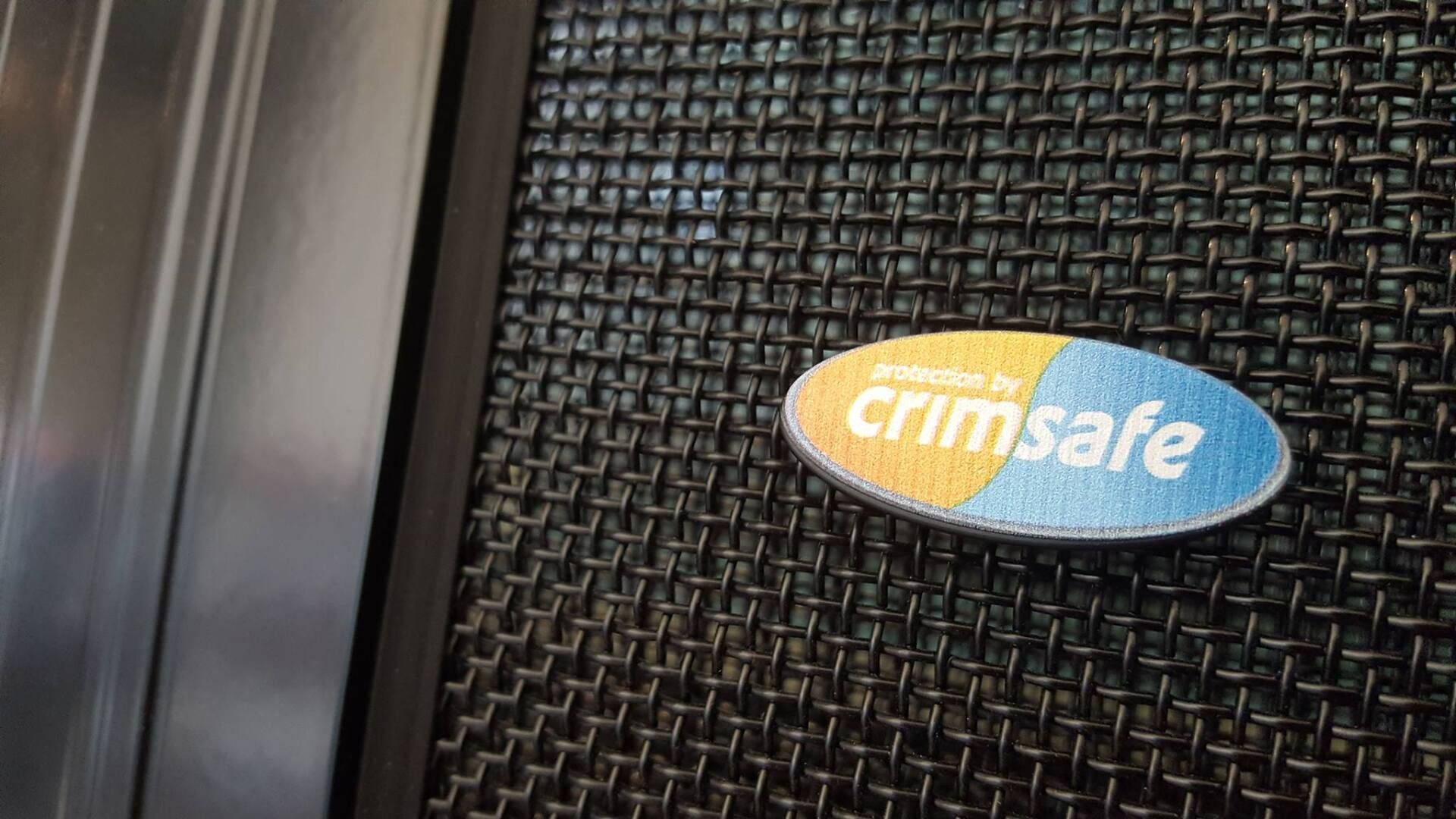 Close-up of a Crimsafe security screen with the logo visible. Black mesh with a blue and yellow oval logo.