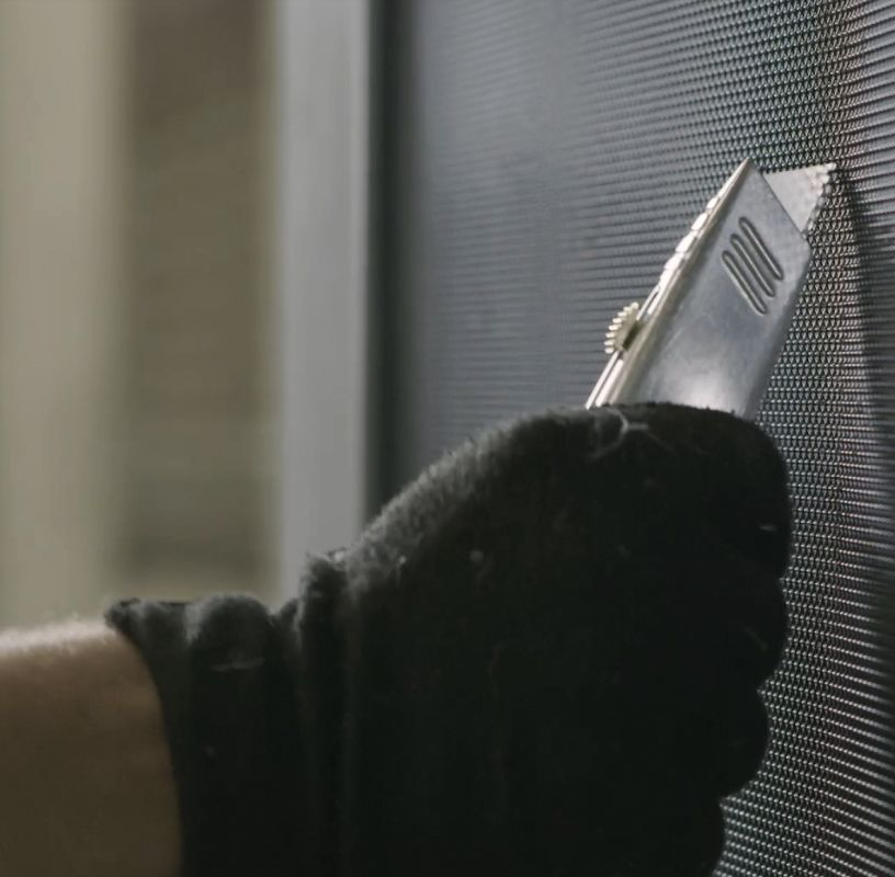 Gloved hand using a utility knife to trim excess screen material from a window or door frame.