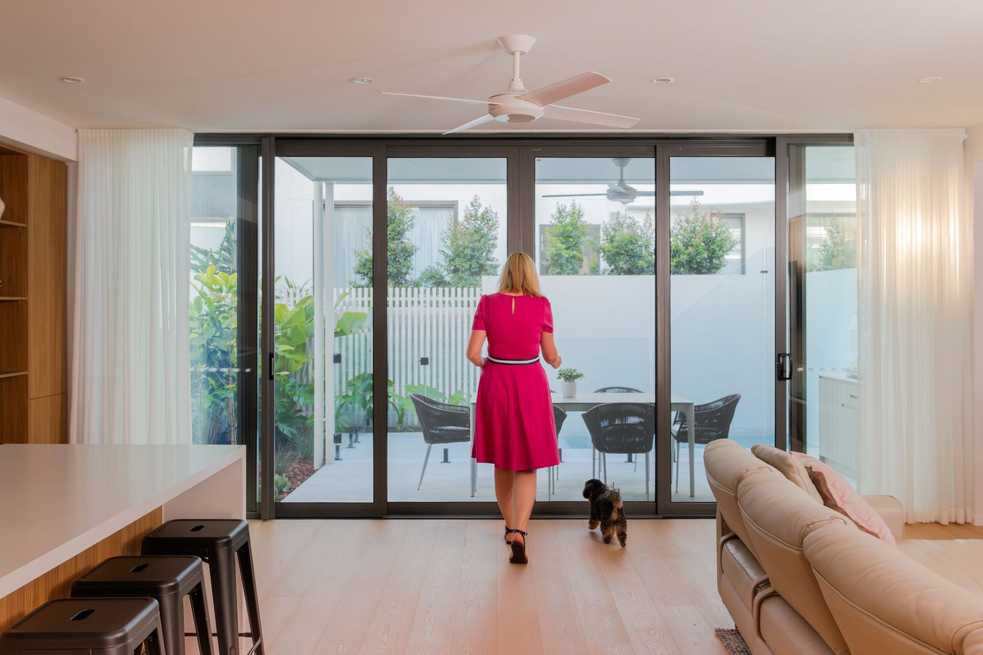 Woman in pink dress walks toward patio, dog follows. Interior view with open sliding doors, neutral décor.
