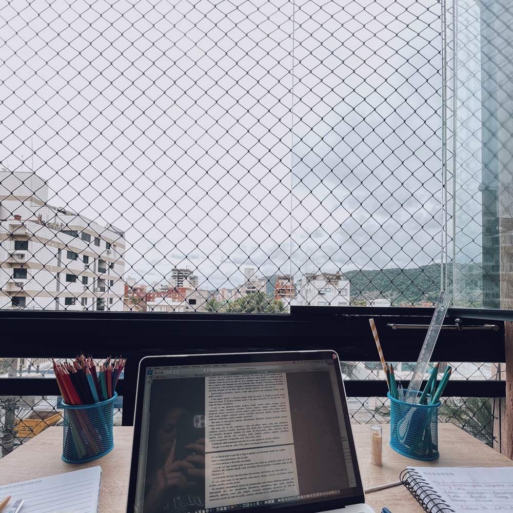 Laptop on a desk by a window with a city view, surrounded by colored pencils and a notebook.