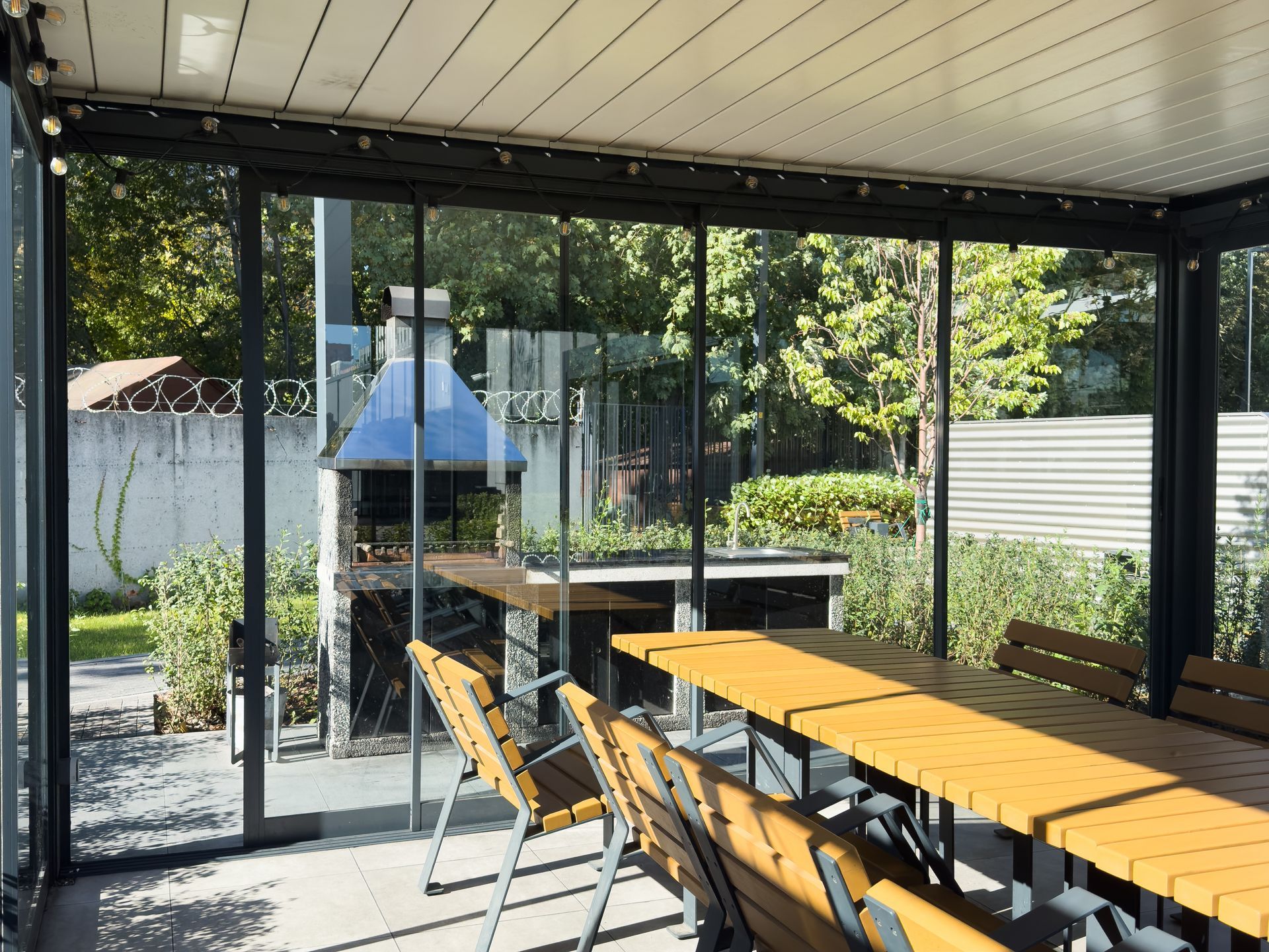 Outdoor dining area with glass walls, wooden table and chairs, overlooking a grill and garden.