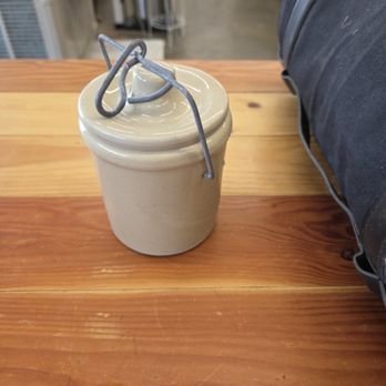 Tan ceramic jar with lid and wire handle, sitting on a wooden surface.