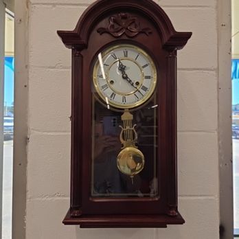Wooden wall clock with a pendulum, Roman numerals, and a gold-colored face, hanging on a white brick wall.
