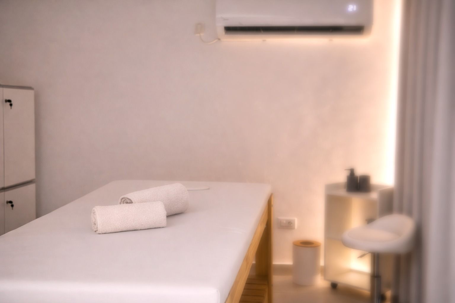 Una camilla de masaje con dos toallas enrolladas en una habitación minimalista, que cuenta con un armario blanco.