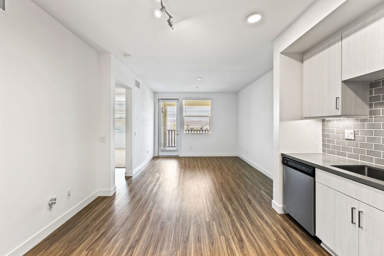 Open-concept living area with wood-style flooring, contemporary kitchen on the right, and balcony door at the far end.