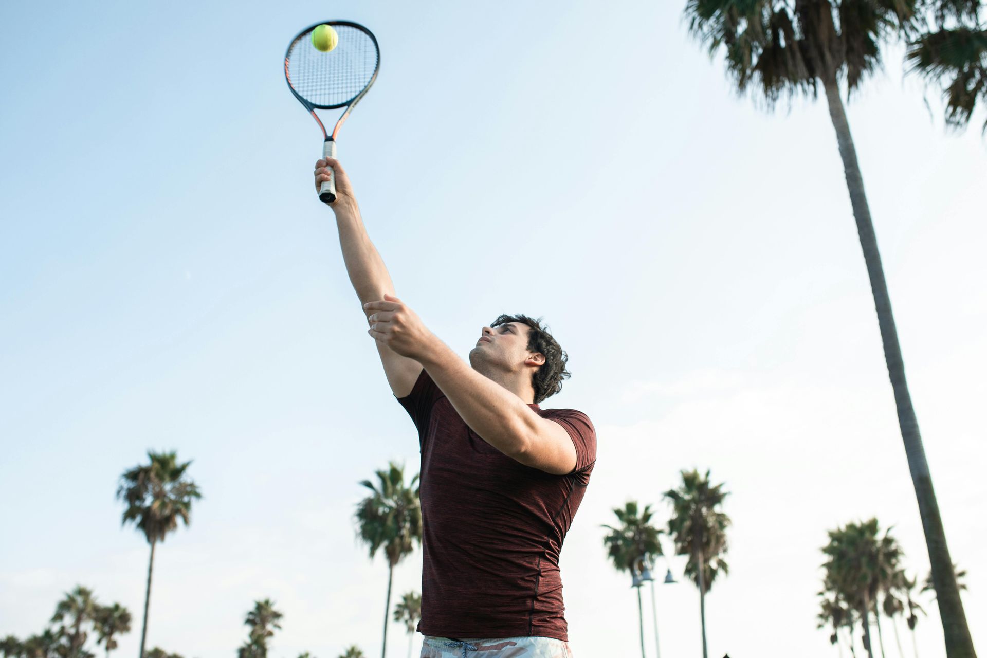 Man serving a tennis ball, arm outstretched, with palm trees and blue sky.