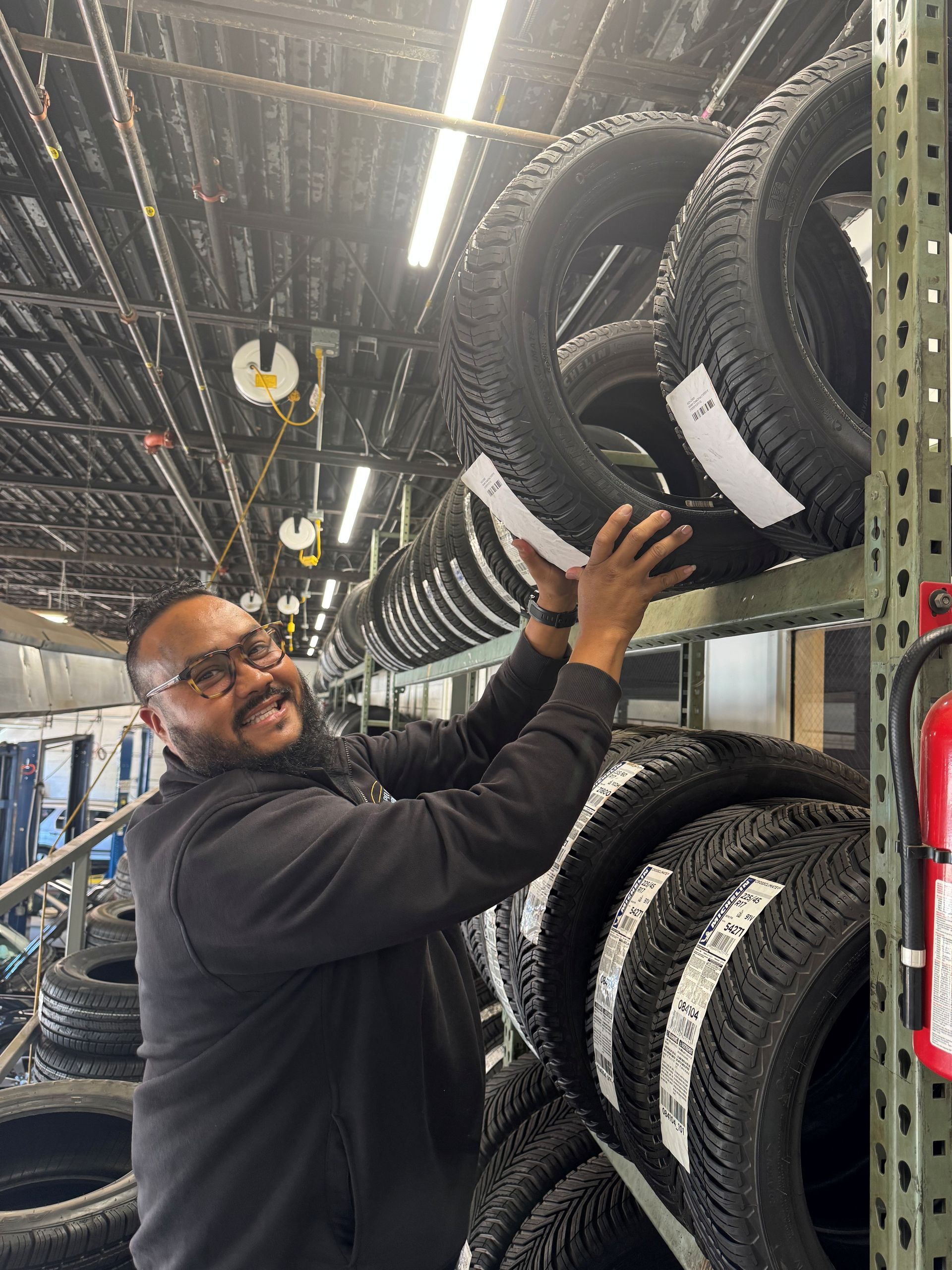 Man reaching for a tire in a warehouse. Tires are stacked on shelves. He is smiling.