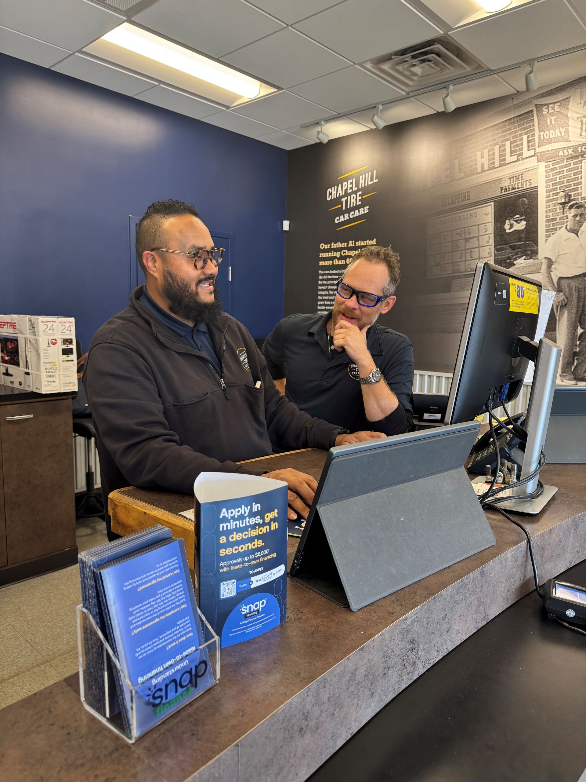 Two people at a desk: one man with glasses looking at a laptop, another at a monitor. Indoors, blue and dark setting.