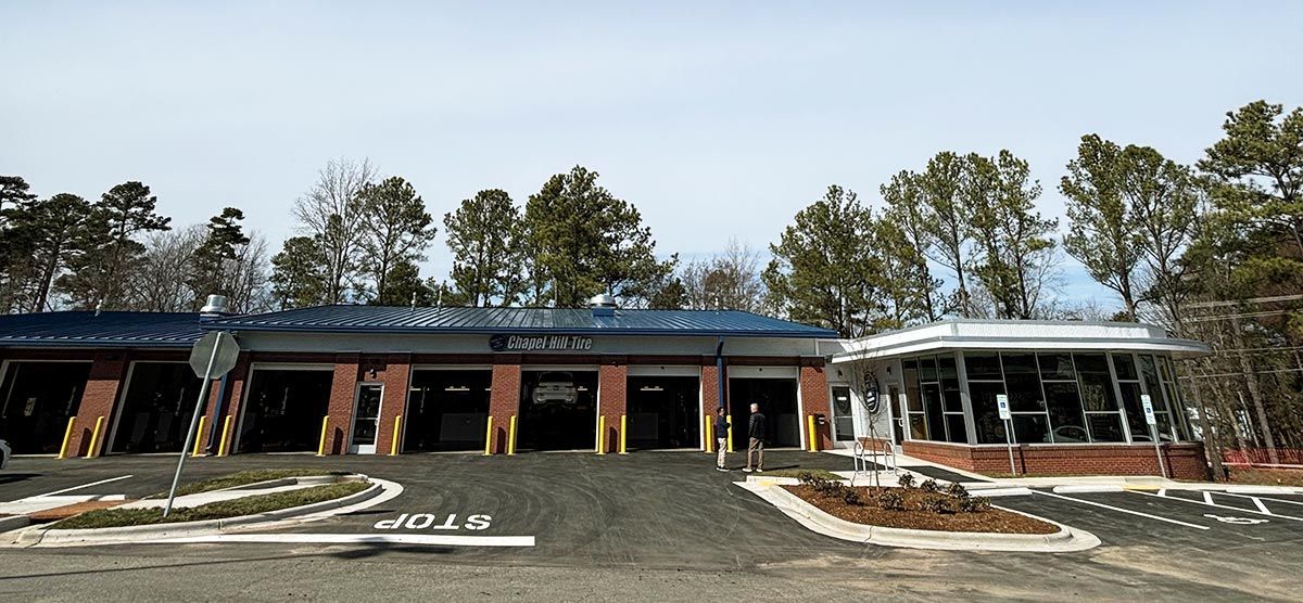 Car repair shop with brick and glass building, gravel driveway, and trees in the background.