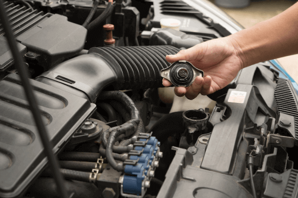 A person is opening the hood of a car and holding a radiator cap.